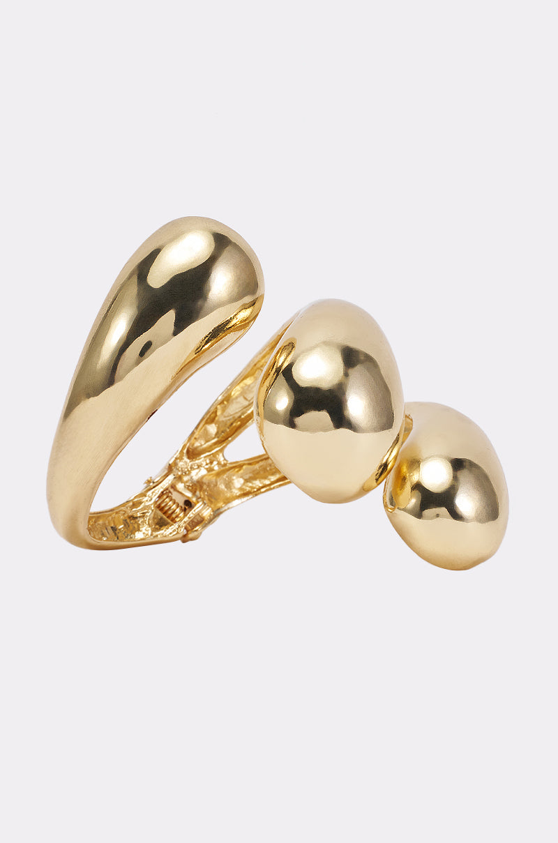 A shiny gold cuff named the CECELIA METAL CUFF IN GOLD, featuring three large, smooth, pebble-like sections on a polished band and set against a simple light background—an ideal statement piece to pair with your favorite chunky gold or sculptural bracelet for a bold, contemporary style.
