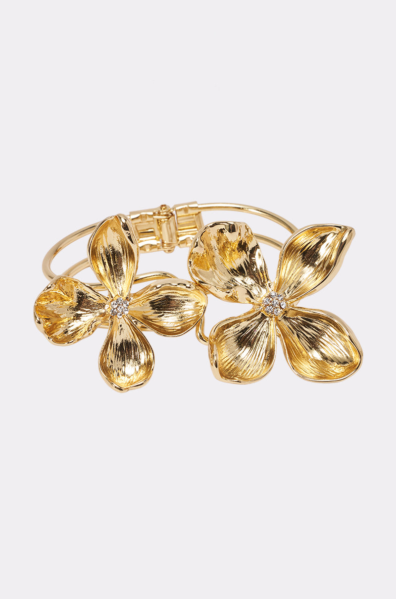 The IN THE GARDEN BRACELET CUFF is a gold cuff adorned with two large textured floral designs, each accented with sparkling rhinestone centers, shown against a white background.