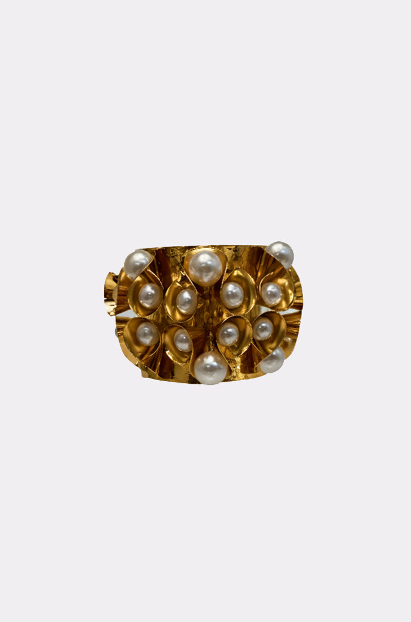 The PEARL PLEASE CUFF is a bold gold-tone bracelet with raised accents and round white pearls, set against a plain light gray background, making it a striking statement accessory.
