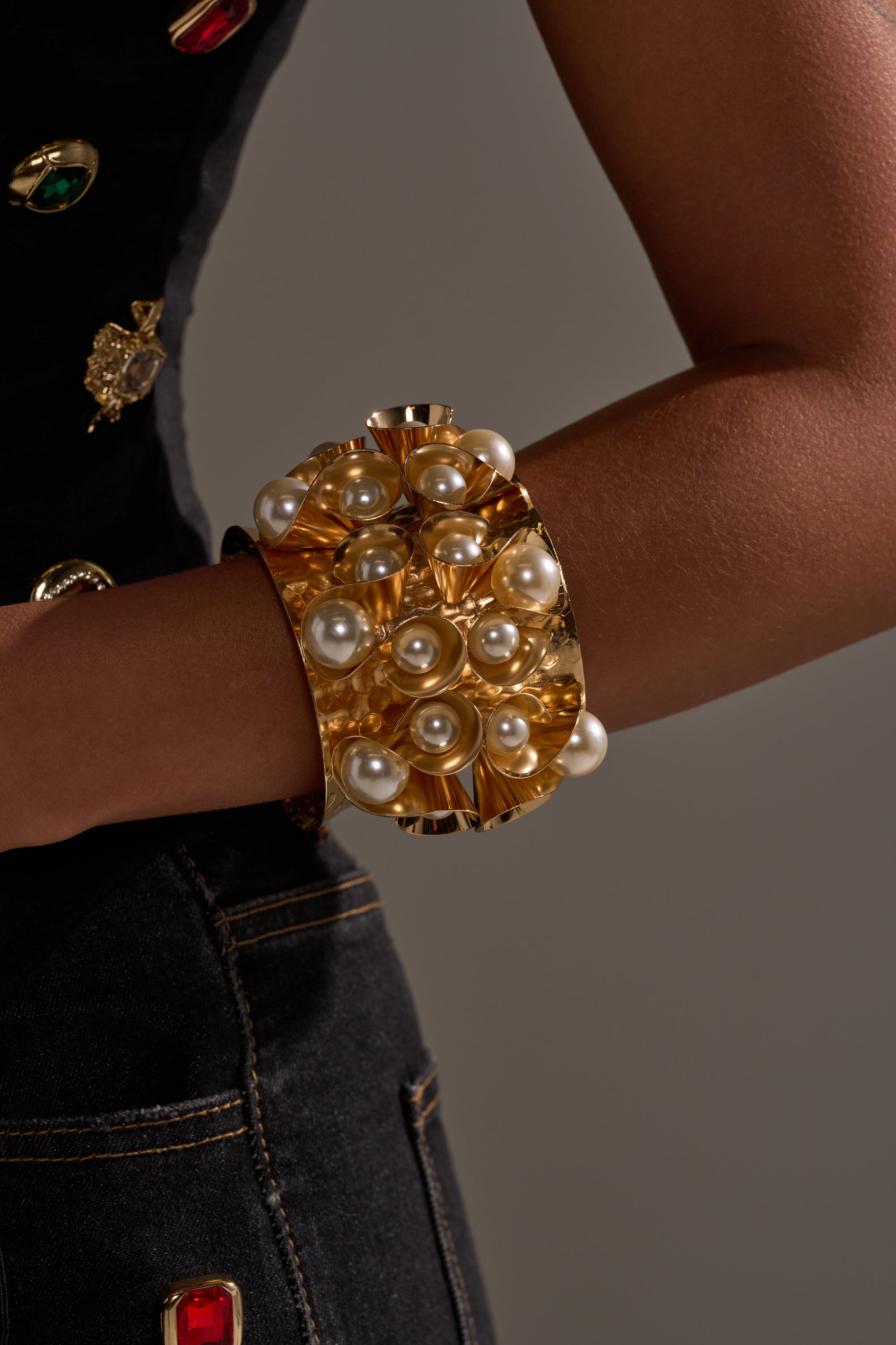 A person wears the PEARL PLEASE CUFF, a bold gold-tone bracelet with clusters of white pearls, paired with a dark sleeveless top and dark denim jeans featuring gold stitching.