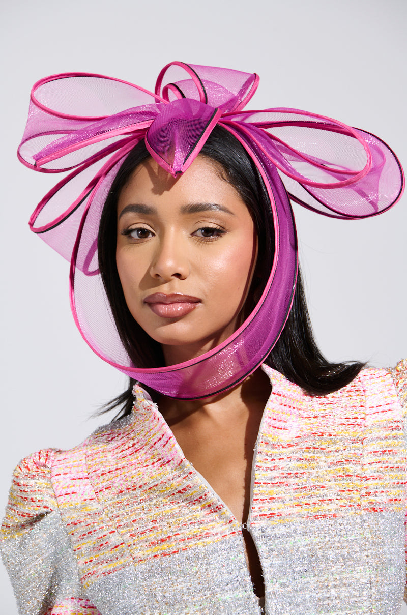 A person with long dark hair wears the TO THE RACES Fascinator in pink, a vibrant Derby Party Accessory with large loops. They're dressed in a textured, multicolored outfit featuring shades of pink, yellow, and white against a plain, light background. 0