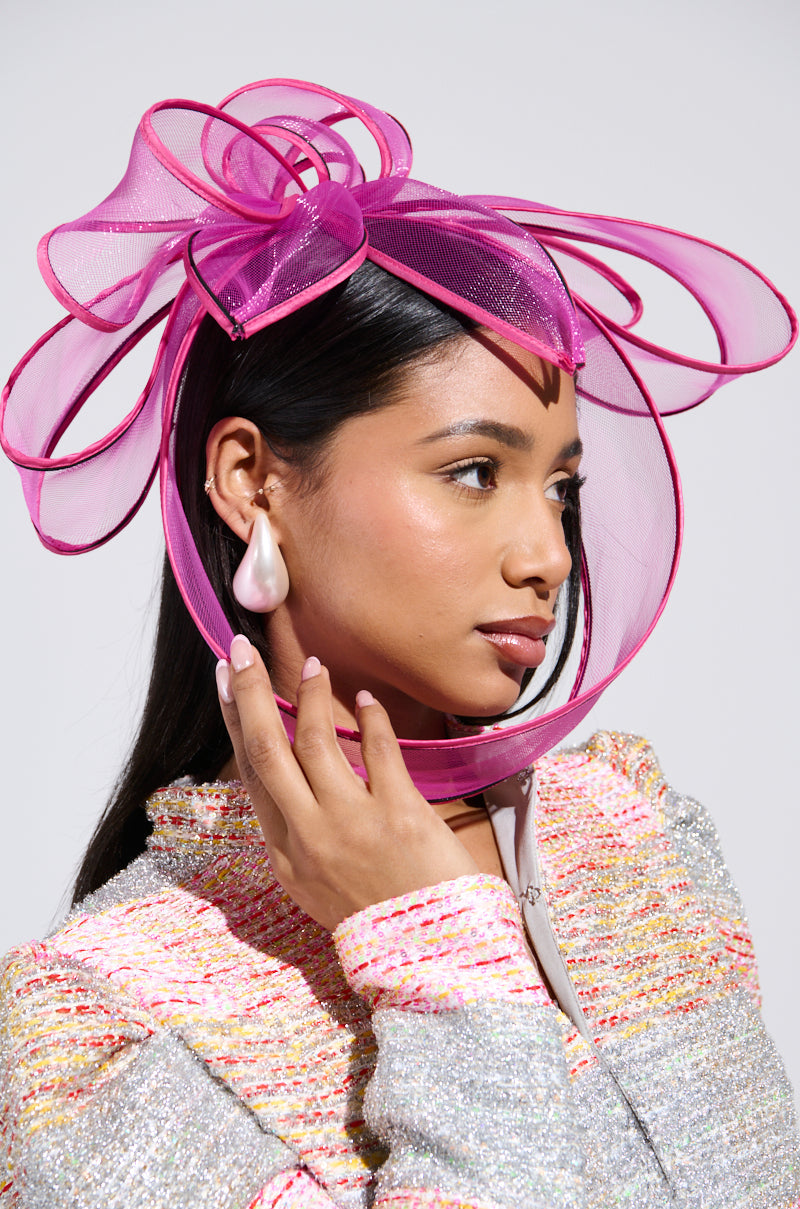 A woman dons a vibrant "To The Races" fascinator in pink with large loops and a pearl earring. Her long dark hair cascades as she touches the fascinator, her outfit textured in multi-colored pastels, creating an ensemble ideal for any Derby party accessory collection. 1