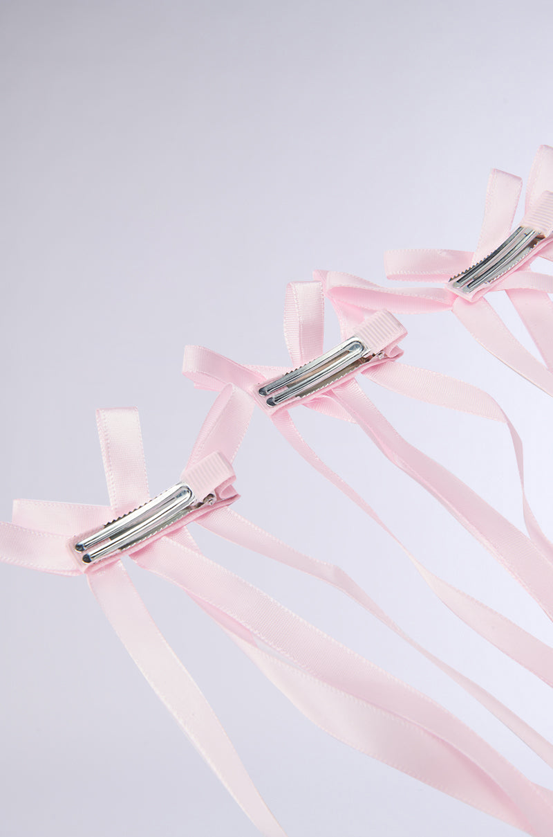The CUTESY HAIR CLIP SET features three pink barrettes with satin ribbons lined up on a light gray background, each adorned with long, flowing ribbons for a delicate and feminine look.