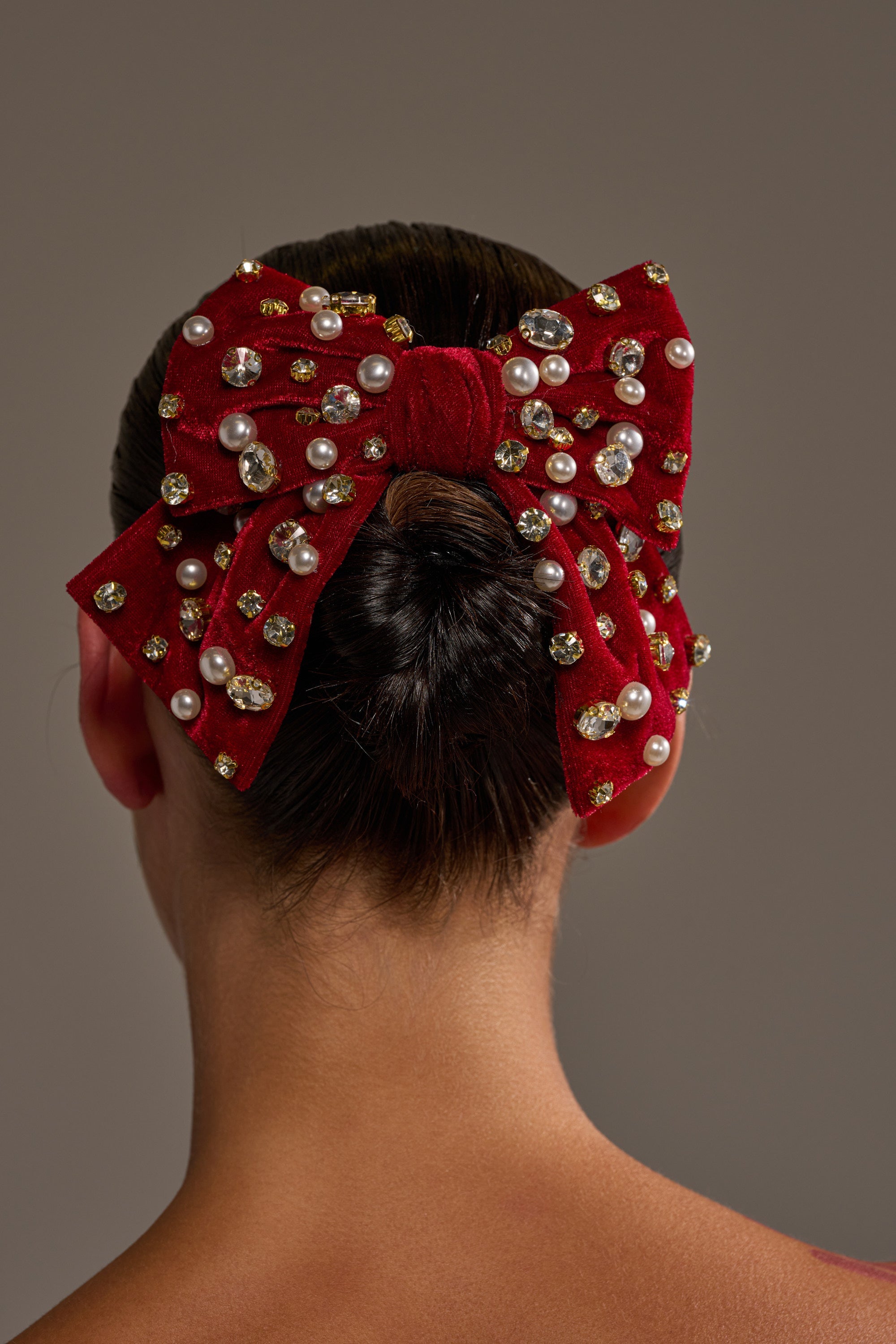 A person with dark hair in a low bun, viewed from behind against a neutral background, wears the JENNI EMBELLISHED HAIR BOW—an oversized velvet accessory adorned with pearls and gold gems. 0