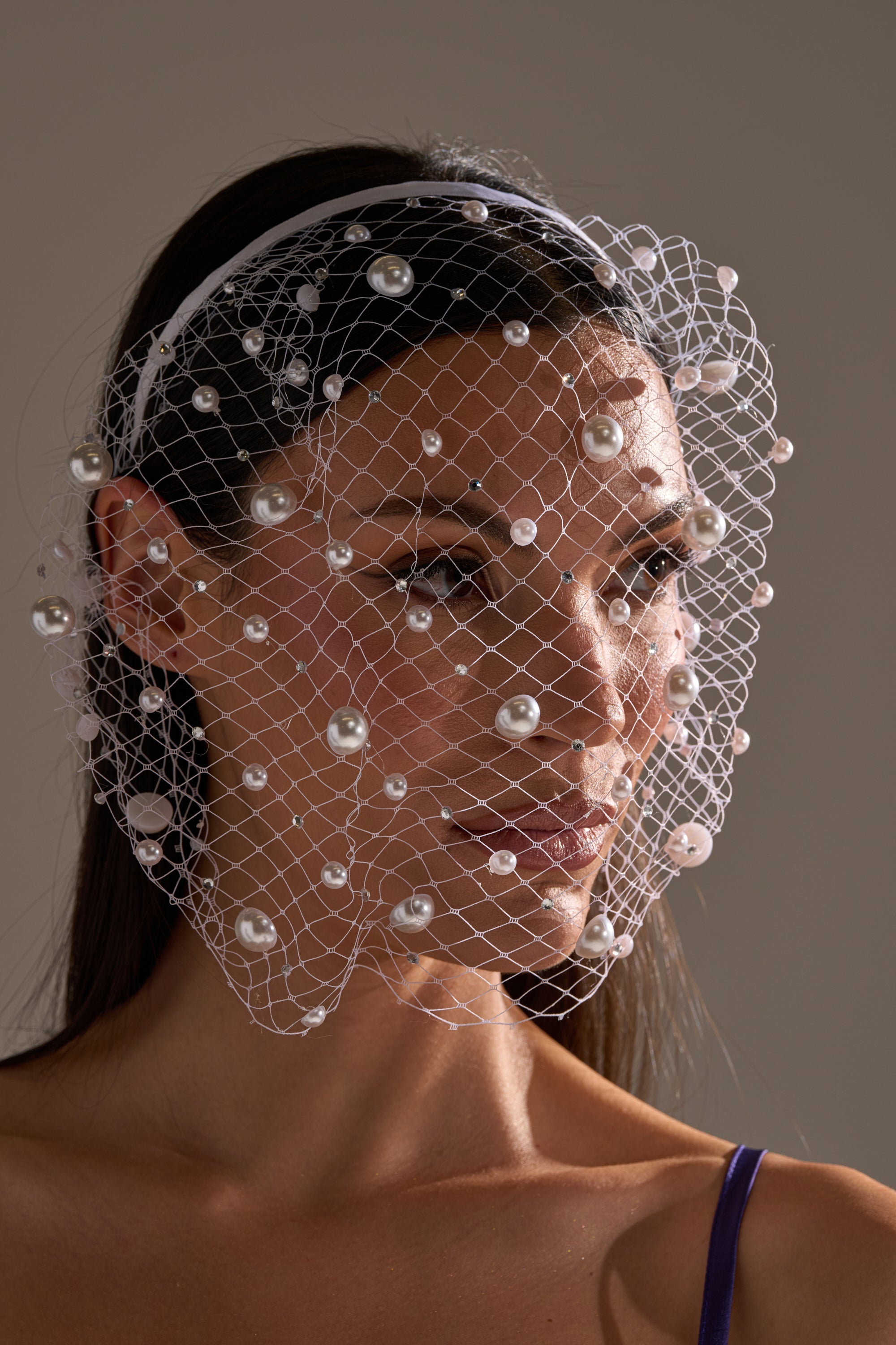A woman with long dark hair models the KYLIE PEARL HEADBAND, styled with a decorative white pearl veil and a purple spaghetti strap top, set against a neutral background. 0