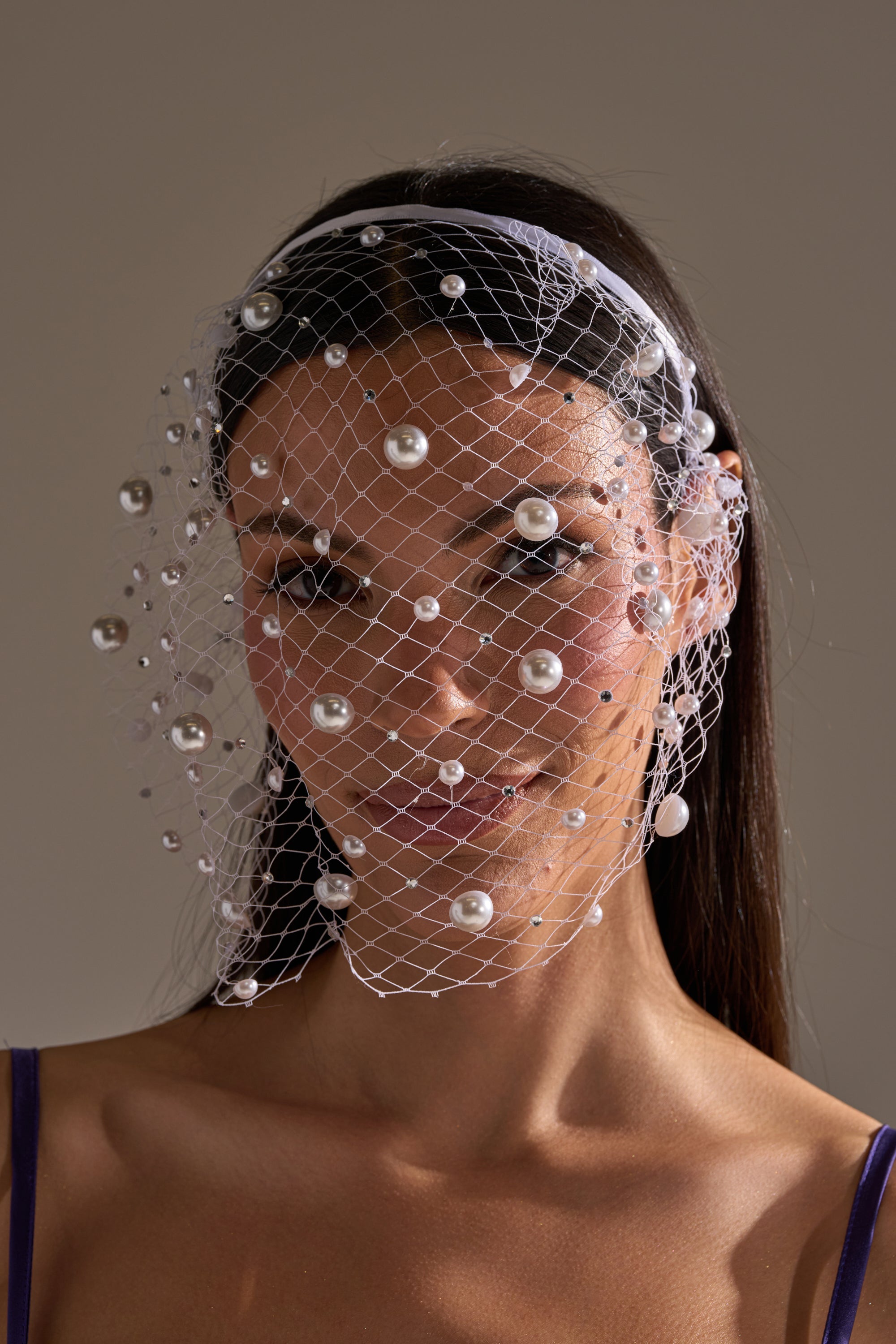A woman with straight dark hair wears the KYLIE PEARL HEADBAND, featuring a white netted veil adorned with pearls over her face. She's dressed in a purple spaghetti strap top against a neutral background. 1