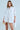A woman poses in the SHE MEANS BUSINESS MINI DRESS, a flowy white button-down with long sleeves crafted from a cotton blend. She holds a gold clutch, stands against a plain light background, and accessorizes with soft waves and a gold necklace.