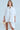 A person in the SHE MEANS BUSINESS MINI DRESS, a flowing white button-down mini dress, stands against a plain background. They hold a beige clutch with long brown hair, gold earrings, and a confident expression. The cotton blend fabric enhances their elegance.