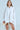 A person stands confidently in the SHE MEANS BUSINESS MINI DRESS, a flowing white shirt dress with an asymmetrical hem made from a cotton blend, holding a small beige clutch. They have long hair and a neutral expression set against a blue background.
