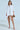 Against a light background, an individual wears the SHE MEANS BUSINESS MINI DRESS, paired with a gold clutch and high-heeled shoes. They have long hair.