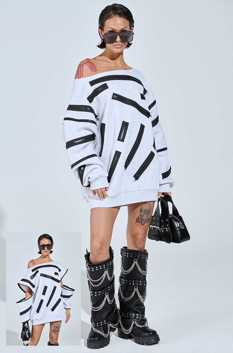 The individual wears a Zip Zap Off-The-Shoulder Sweatshirt Dress in Grey Multi, large black sunglasses, and black boots with silver chains. They're holding a chic handbag, with tattoos on their legs and shoulder. A smaller photo showcases the same captivating pose.