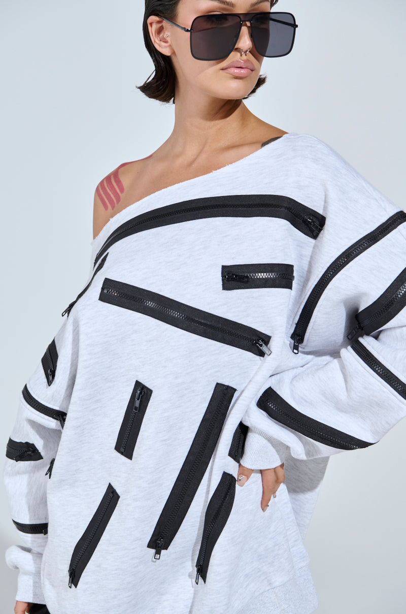 A person with short hair wears large square sunglasses and the ZIP ZAP OFF THE SHOULDER SWEATSHIRT DRESS-GREY MULTI, which features dynamic zipper detailing. The dress casually exposes a pink tattoo as it falls off the shoulder.