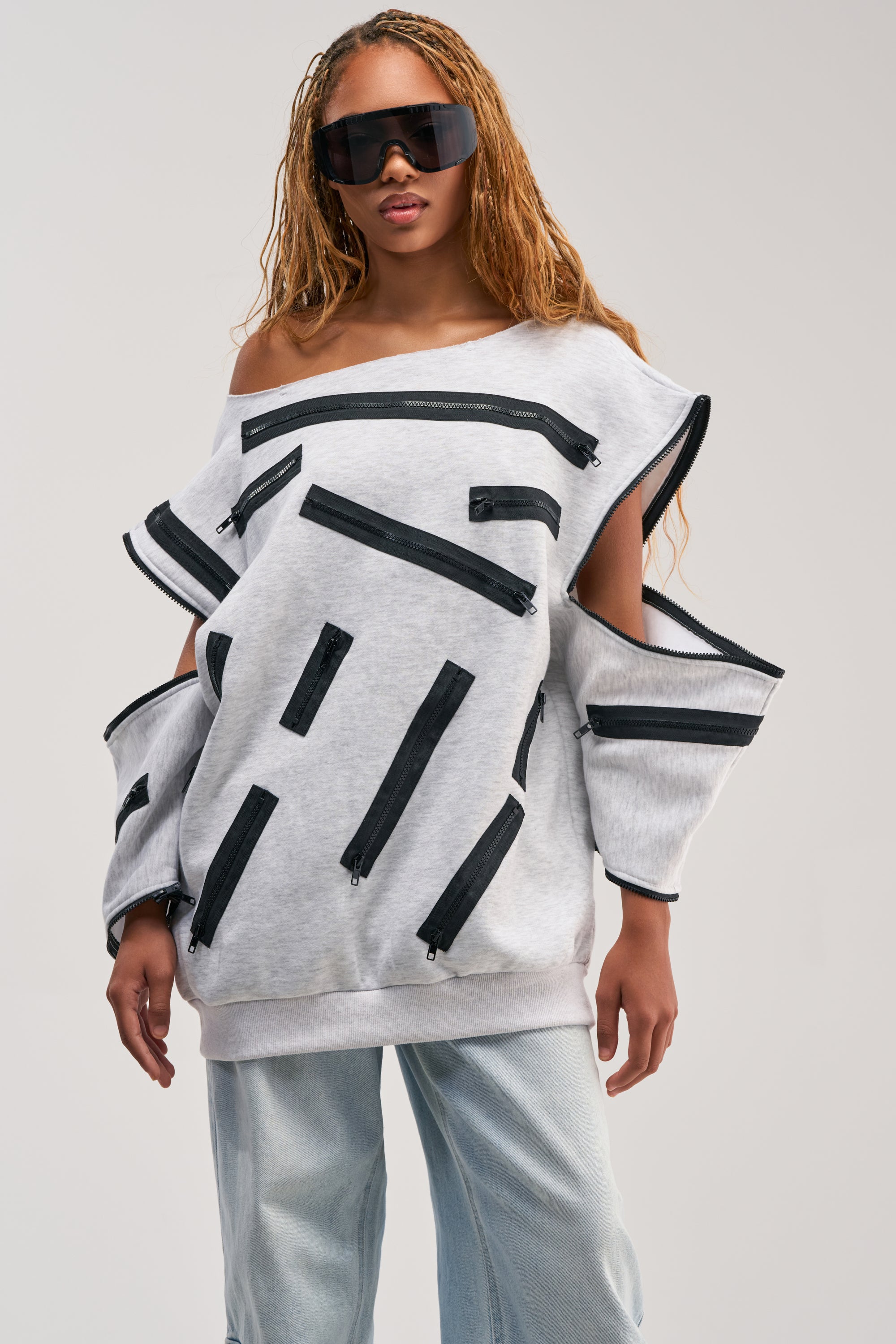Woman wearing the ZIP ZAP OFF THE SHOULDER SWEATSHIRT DRESS-GREY MULTI—an oversized light gray streetwear dress with multiple black zippers—styled with light blue jeans, large black sunglasses, and long braided hair, standing against a plain background.