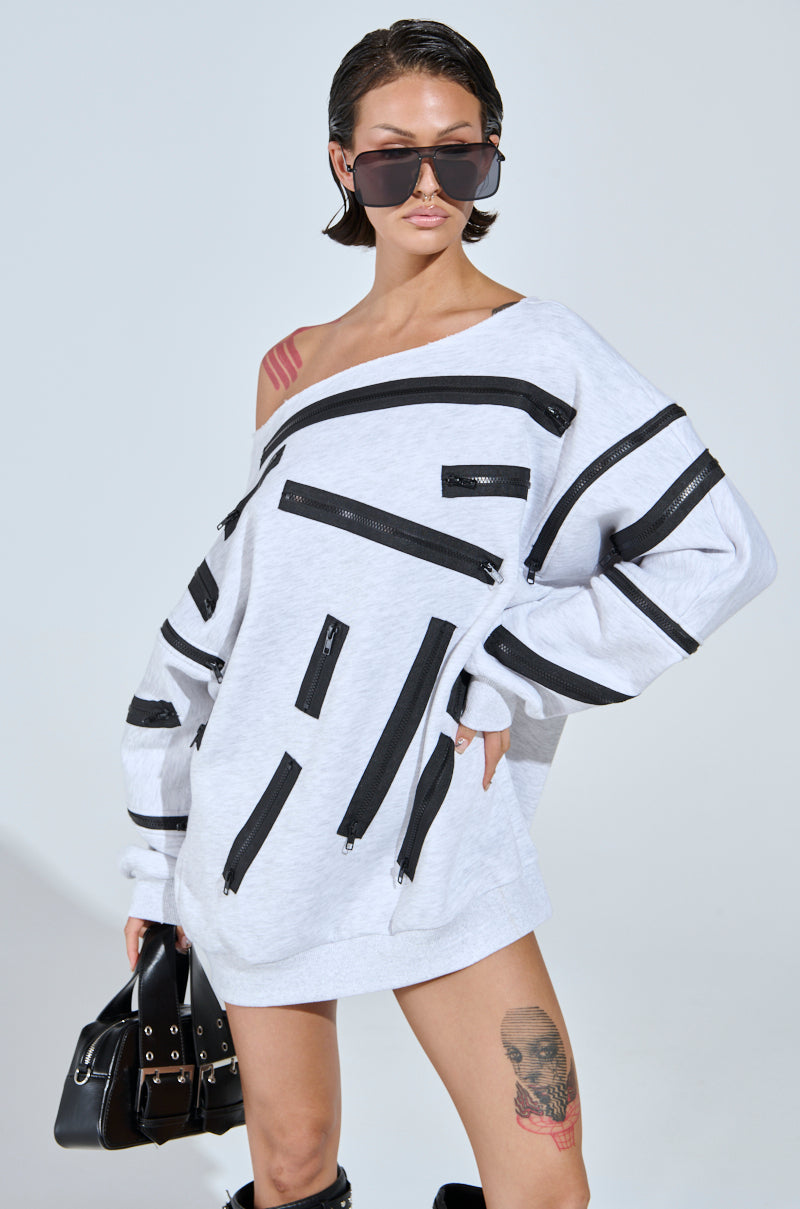 A person with short hair wears large sunglasses and an oversized, off-the-shoulder sweatshirt dress in grey multi from the ZIP ZAP collection, featuring black zippered stripes. They accessorize with a black bag, knee-high boots, and display a colorful tattoo on their left thigh.