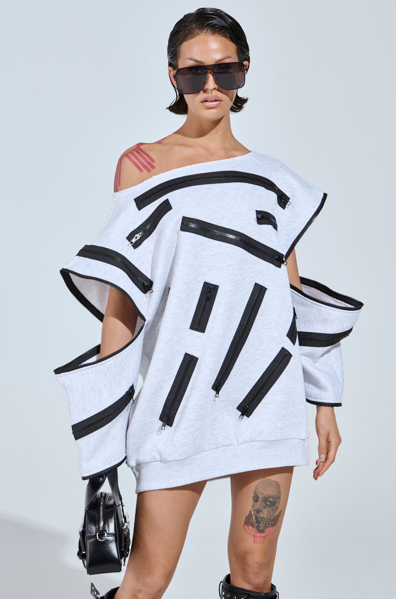 Wearing a grey multi sweat dress named "ZIP ZAP OFF THE SHOULDER SWEATSHIRT DRESS," they exude confidence with bold black zipper detailing. Sporting short dark hair, oversized sunglasses, tattoos of a red stripe on the shoulder, and a face on the leg.
