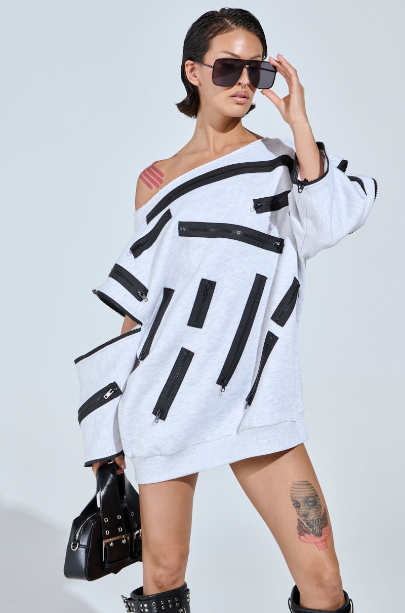 A person in an oversized off-the-shoulder sweatshirt dress poses confidently with large sunglasses, a small black handbag, and knee-high studded boots. A visible tattoo adorns their left thigh.