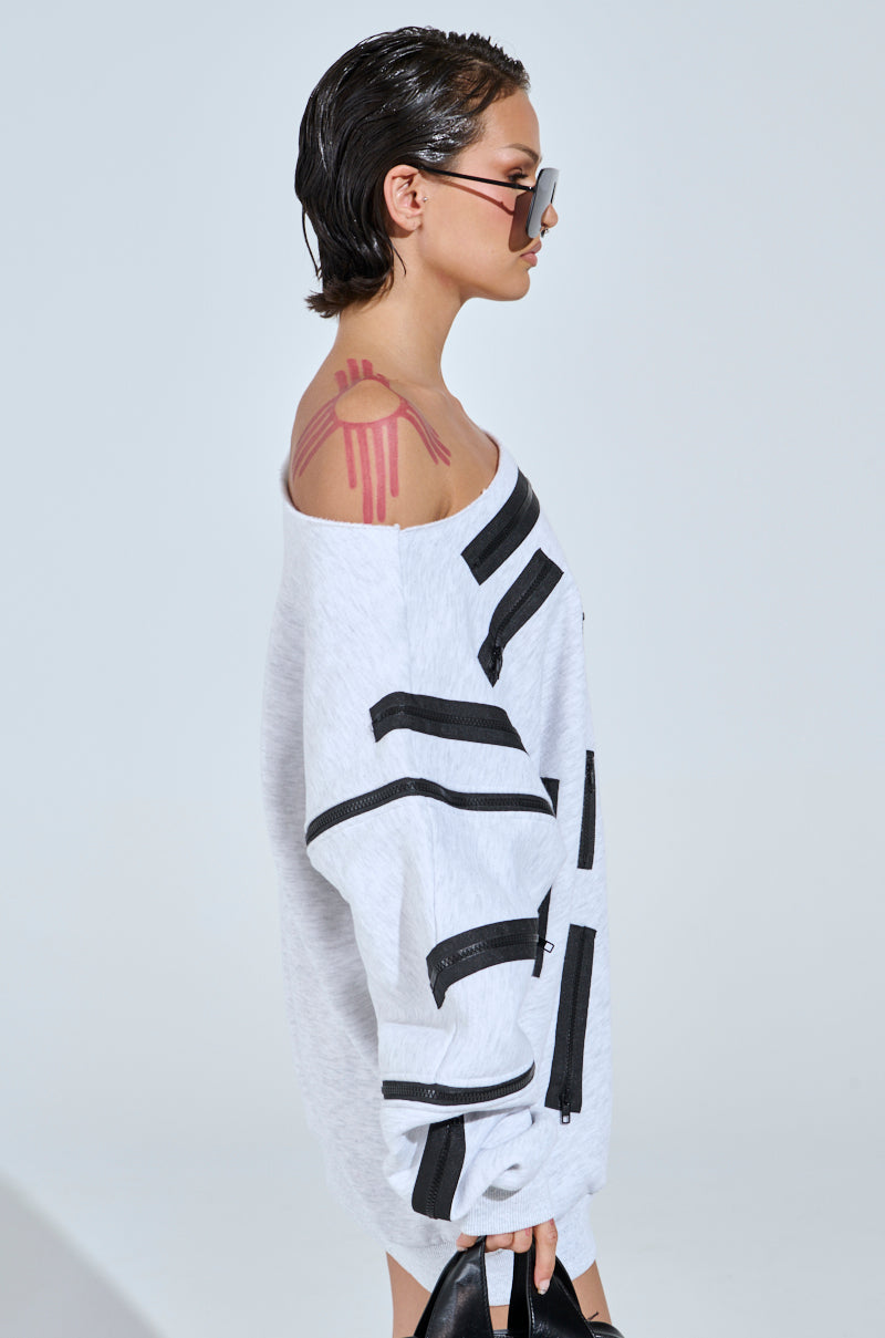 A person with slicked-back hair and sunglasses wears the ZIP ZAP OFF THE SHOULDER SWEATSHIRT DRESS-GREY MULTI with zipper sleeve detailing. A red abstract tattoo on their right shoulder enhances the look as they hold black shoes, set against a plain light gray backdrop.