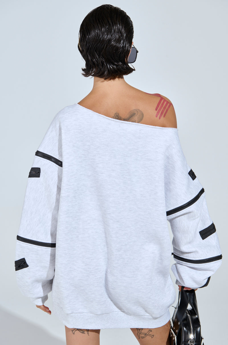 A person with short, dark hair is seen from the back wearing the "ZIP ZAP OFF THE SHOULDER SWEATSHIRT DRESS-GREY MULTI." This loose-fitting dress has black stripe details and trendy zipper sleeve accents. They have a shiny black bag and an upper back tattoo against a plain light gray background.