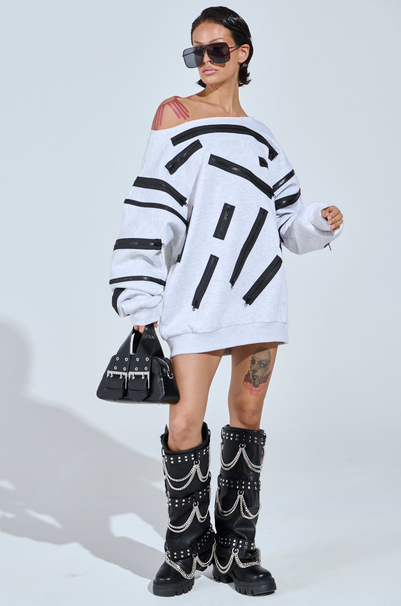 Wearing the ZIP ZAP Off-the-Shoulder Sweatshirt Dress in Grey Multi with black stripe designs, paired with black sunglasses and chunky chain-studded boots, and holding a small black handbag against a plain light grey backdrop.