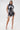 A woman stands confidently in the ALL ABOUT IT LACE UP MINI DRESS IN DENIM, featuring bold white corset-style details. She pairs it with silver high heels and a matching bag, her long dark hair contrasting the plain white background.
