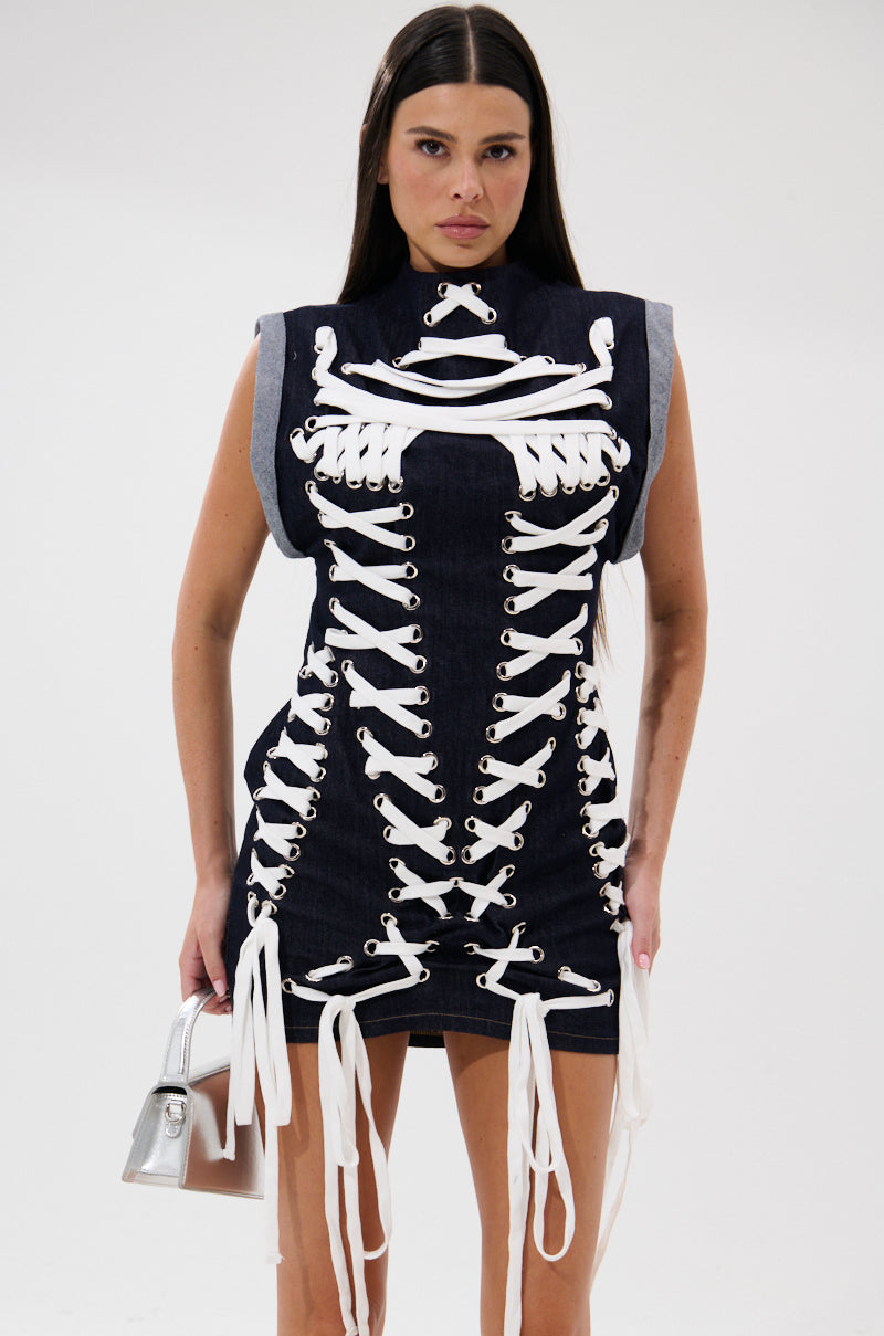 A woman in the ALL ABOUT IT LACE UP MINI DRESS IN DENIM, featuring white skeletal-style details, stands against a plain background holding a small metallic handbag. 2