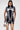 A woman in the ALL ABOUT IT LACE UP MINI DRESS IN DENIM, featuring white skeletal-style details, stands against a plain background holding a small metallic handbag.