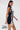 A woman with long brown hair models the ALL ABOUT IT LACE UP MINI DRESS IN DENIM, a sleeveless mini dress featuring white lace-up details on the front and sides. She carries a silver handbag and poses against a plain white background.