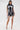 A woman poses against a white background in the ALL ABOUT IT LACE UP MINI DRESS IN DENIM, featuring bold corset-inspired details. She accessorizes with a small silver handbag and pointed silver high heels.