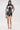 A woman poses against a white background in the ALL ABOUT IT LACE UP MINI DRESS IN DENIM, featuring lace-up details. She accessorizes with a small silver bag and pointed silver heels.