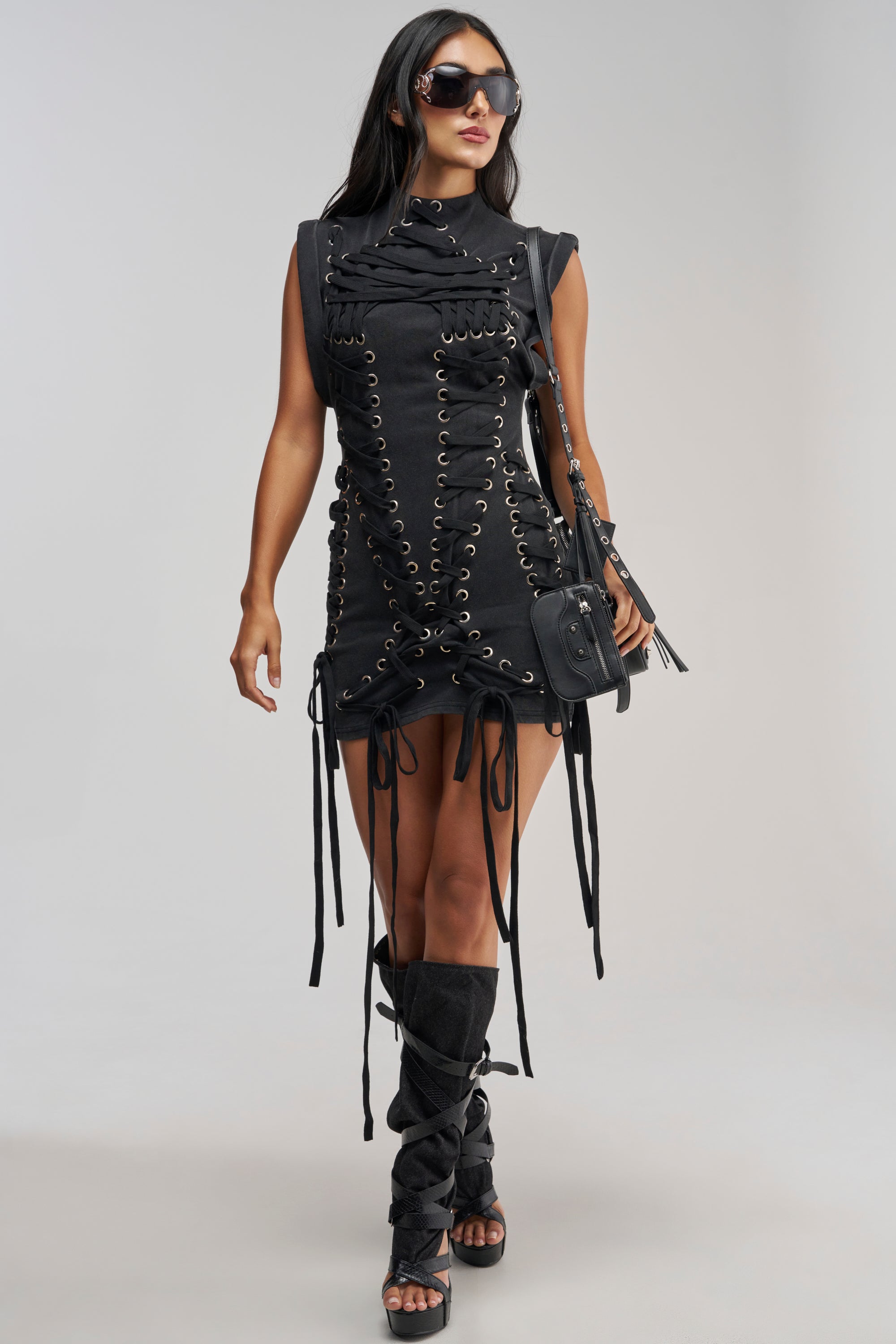 A woman poses confidently against a plain background wearing the BANKS LACE UP MOCK NECK KNIT MINI DRESS, styled with matching lace-up knee-high sandals, black sunglasses, and a black crossbody bag.