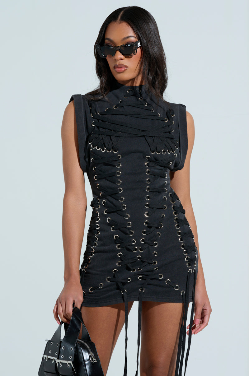A woman in the chic BANKS LACE UP MOCK NECK KNIT MINI DRESS stands against a plain background. Her long dark hair complements her cat-eye sunglasses while she holds a stylish black handbag with metal accents.