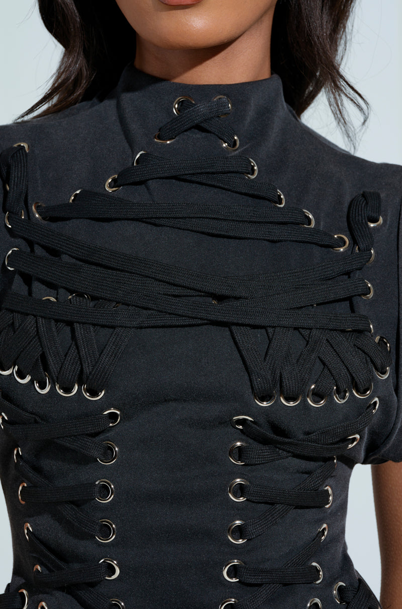 The BANKS LACE UP MOCK NECK KNIT MINI DRESS is worn by a person, showcasing black cotton blend with intricate criss-cross lacing and metal eyelets on the chest and shoulders, creating a textured, layered look. The background is out of focus.