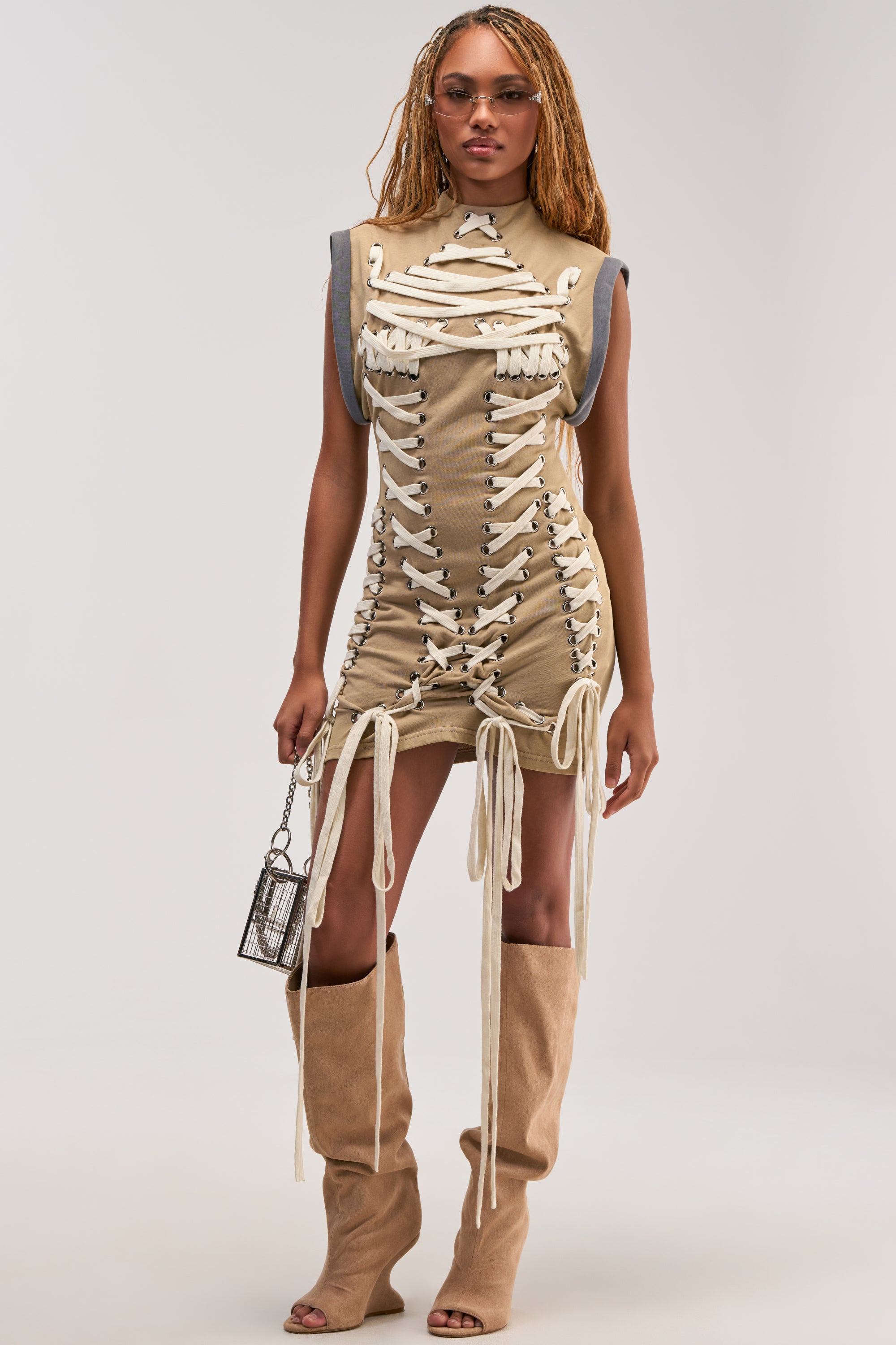 Wearing the BANKS LACE UP MOCK NECK KNIT MINI DRESS IN DESERT, a person with long braids styles their look with tan over-the-knee boots, sunglasses, and a small silver bag, standing against a plain light background for a utility-chic vibe.