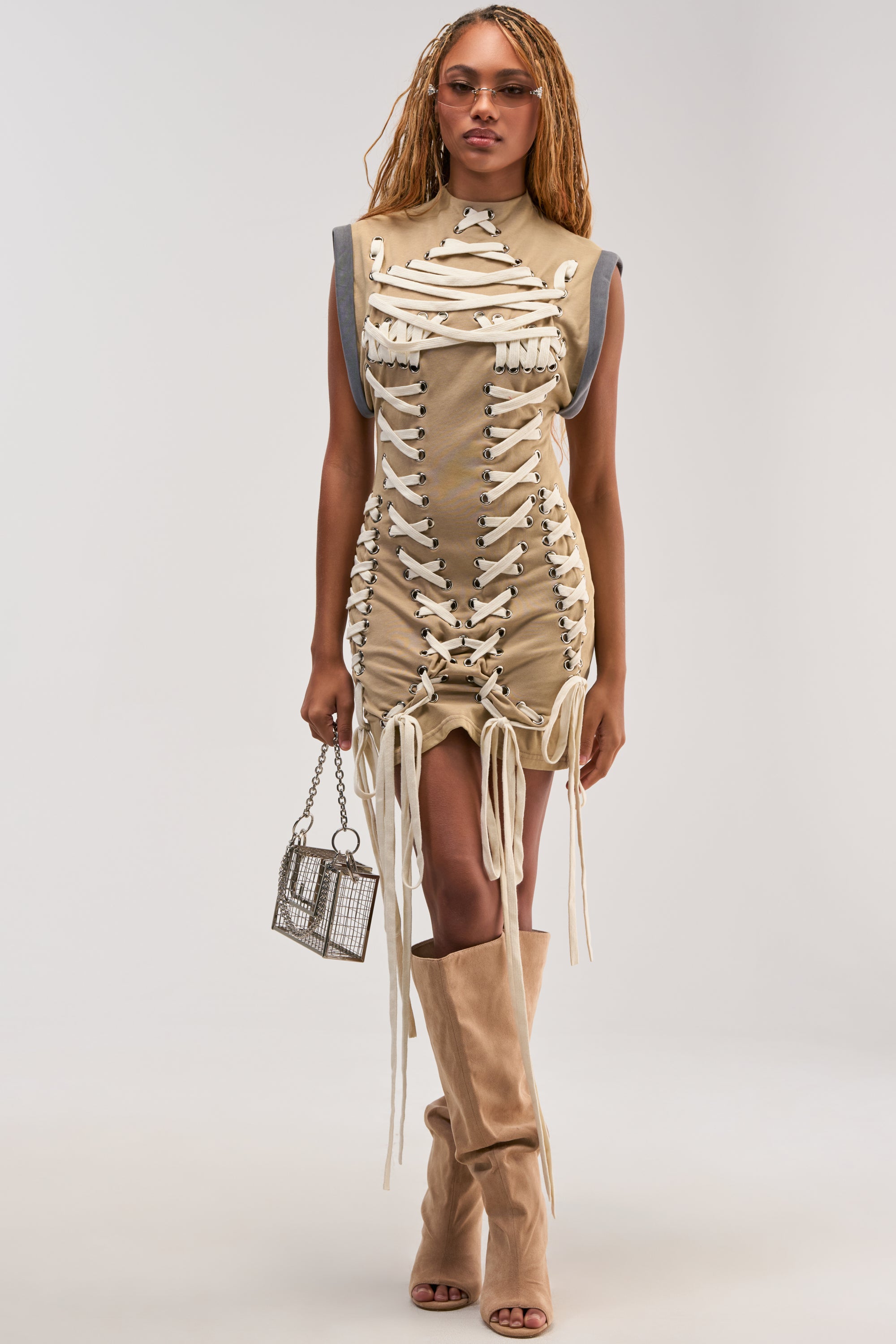 Wearing the BANKS LACE UP MOCK NECK KNIT MINI DRESS IN DESERT, a woman pairs the khaki mini dress with white laces with tan open-toe knee-high boots, sunglasses, and a small silver bag as she stands against a plain background, effortlessly channeling a utility-chic vibe.