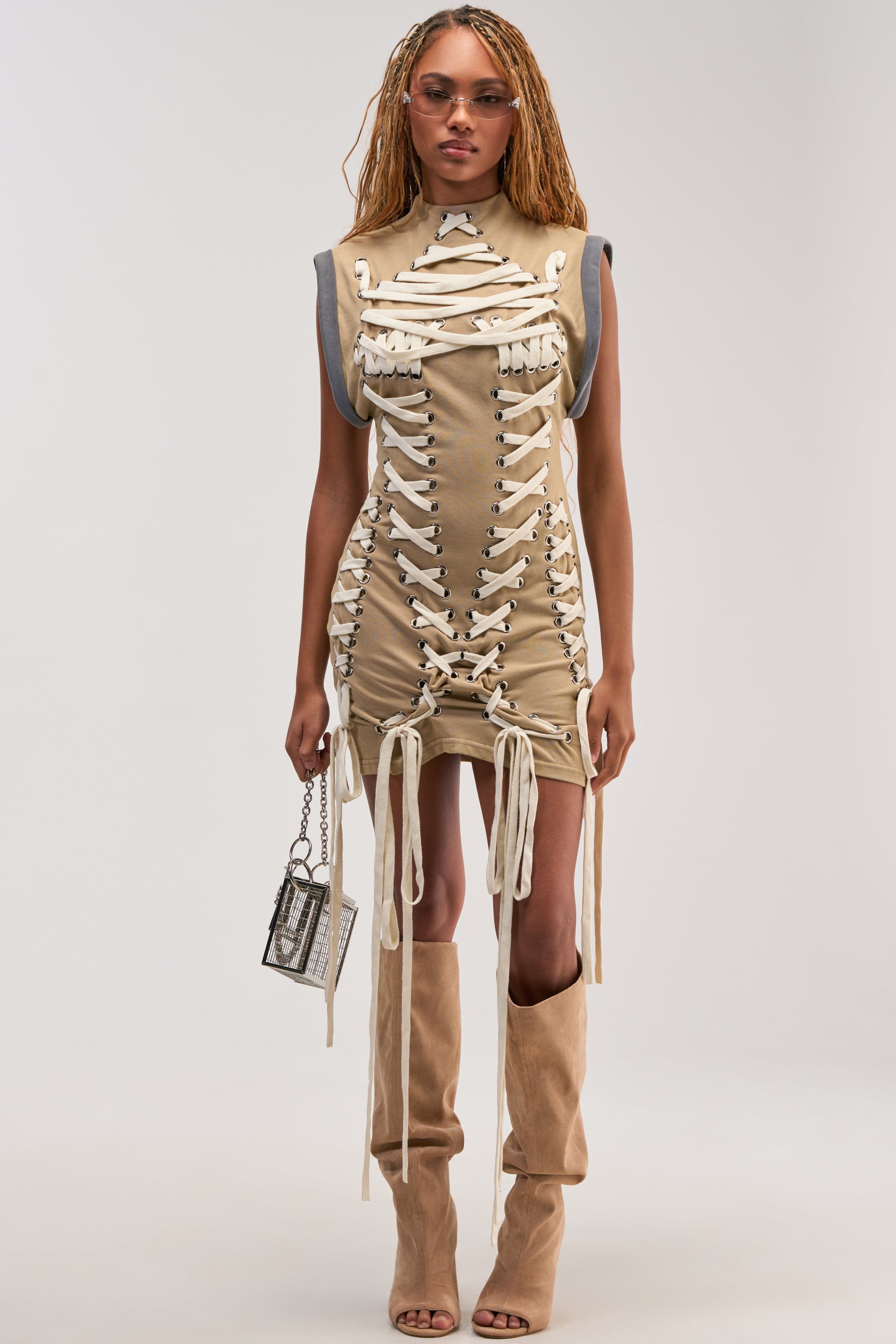 Wearing the BANKS Lace Up Mock Neck Knit Mini Dress in Desert, a woman pairs her look with tan open-toe boots, sunglasses, and a small handbag, standing confidently against a plain background and showcasing an effortless utility-chic style.