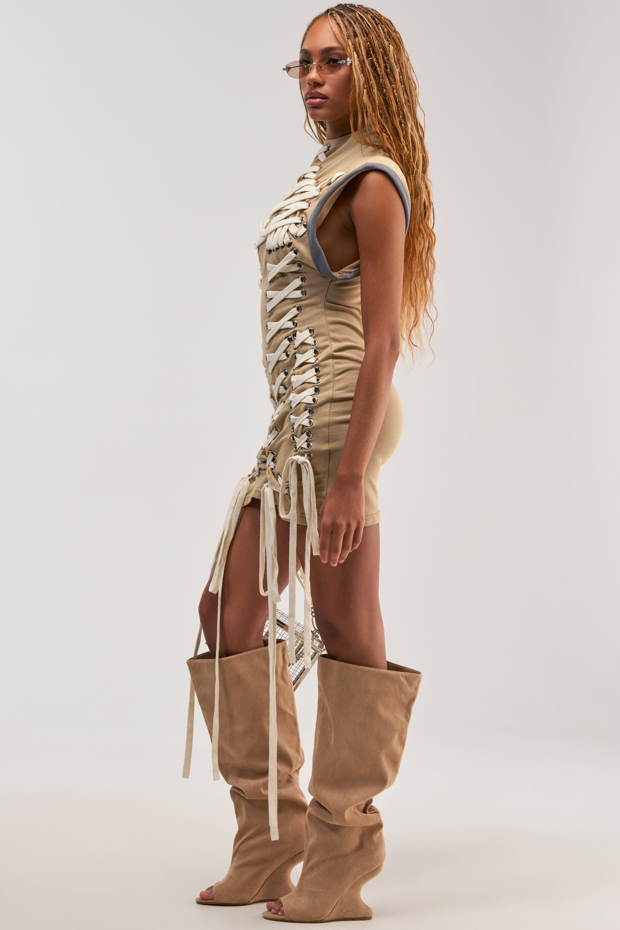 Woman in the BANKS Lace Up Mock Neck Knit Mini Dress in Desert, paired with oversized tan slouchy boots, stands sideways against a plain white background. Sporting long braided hair and sunglasses, she exudes an effortless utility-chic vibe.