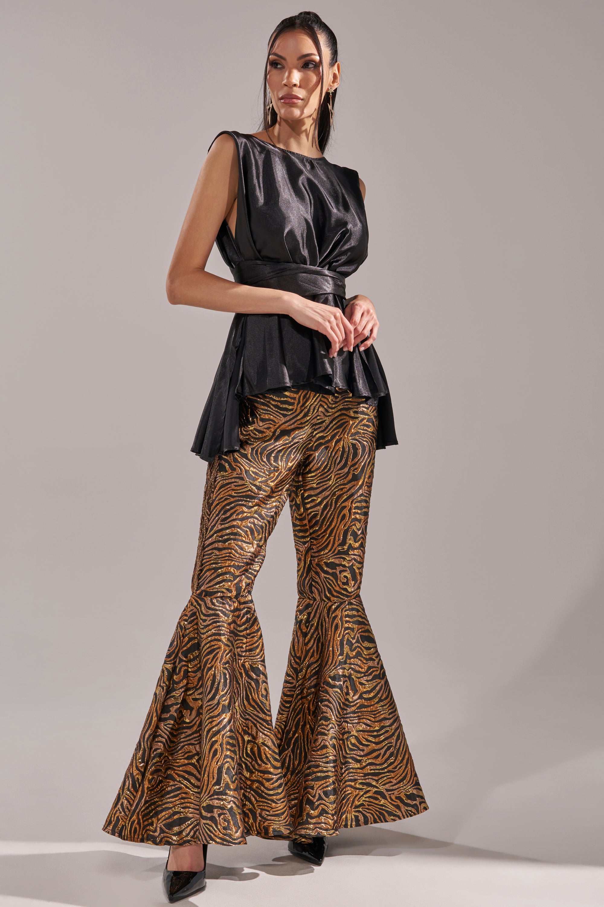 A woman poses against a light background wearing the GET IN MOTION BROCADE FLARE TROUSER, featuring a bold gold and black tiger print, with a sleeveless black belted top and black pointed-toe shoes. 1