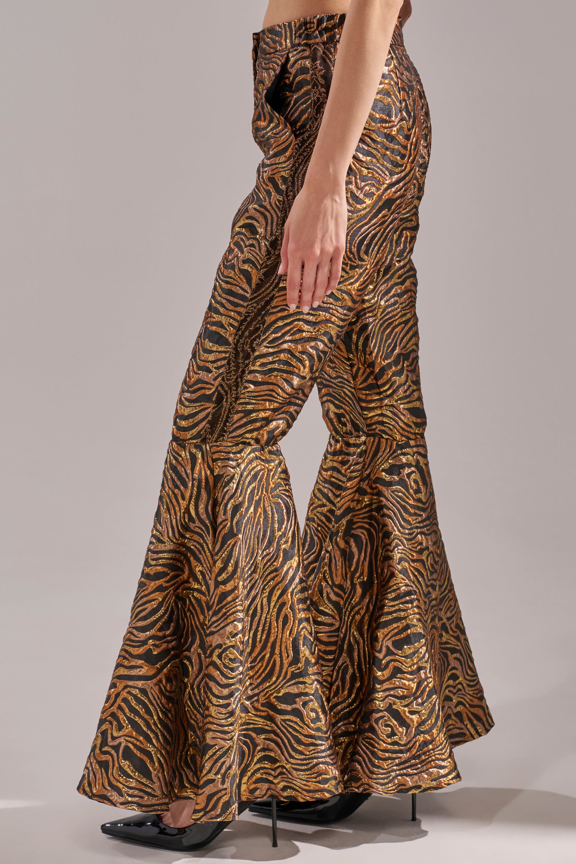 A person shown from the side against a plain background wears the GET IN MOTION BROCADE FLARE TROUSER in metallic gold and black zebra print, paired with black pointed-toe heels. 4