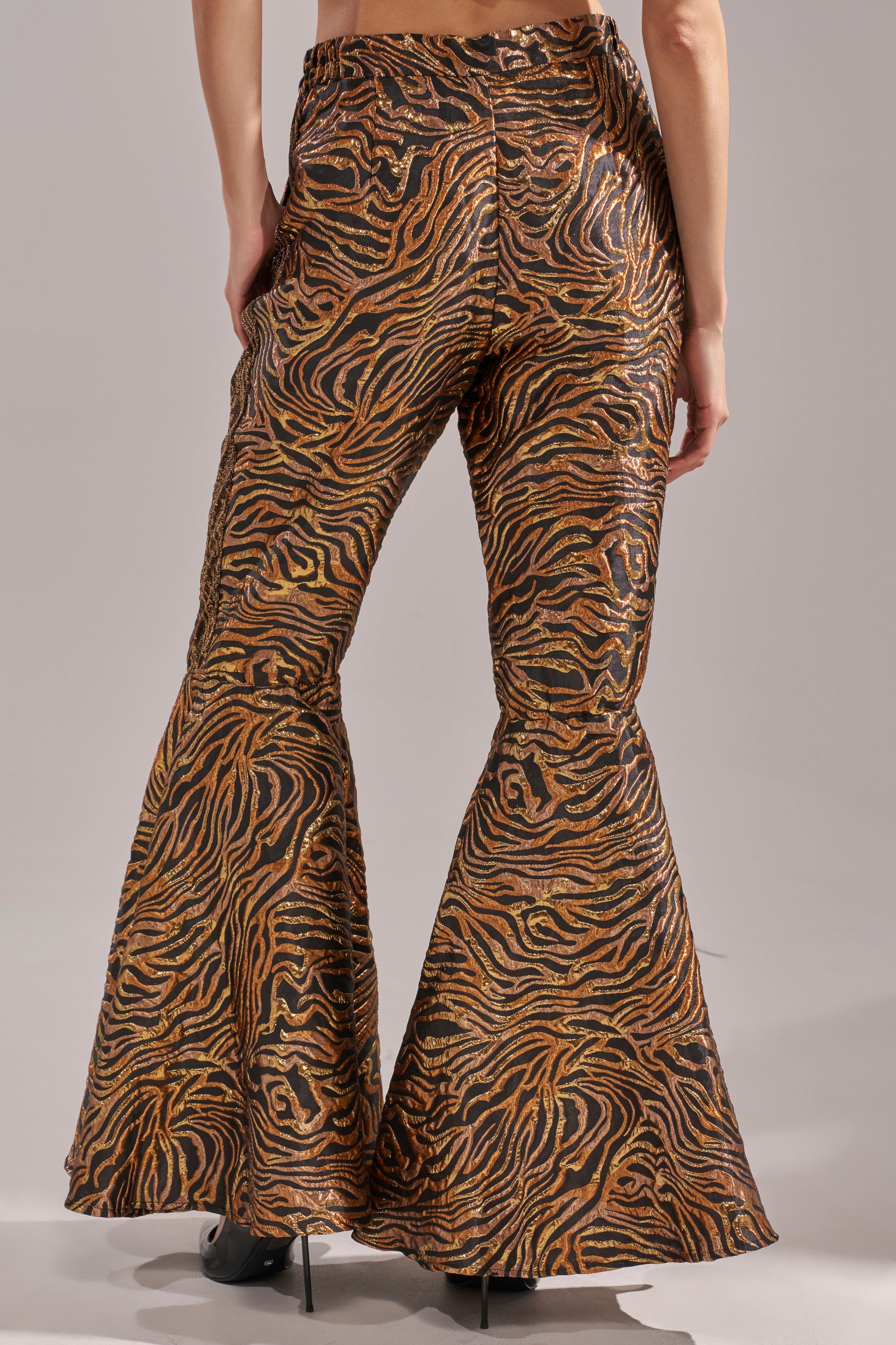 Wearing the GET IN MOTION BROCADE FLARE TROUSER, featuring a metallic gold, brown, and black tiger stripe pattern, a person stands with their back to the camera against a plain background. 5