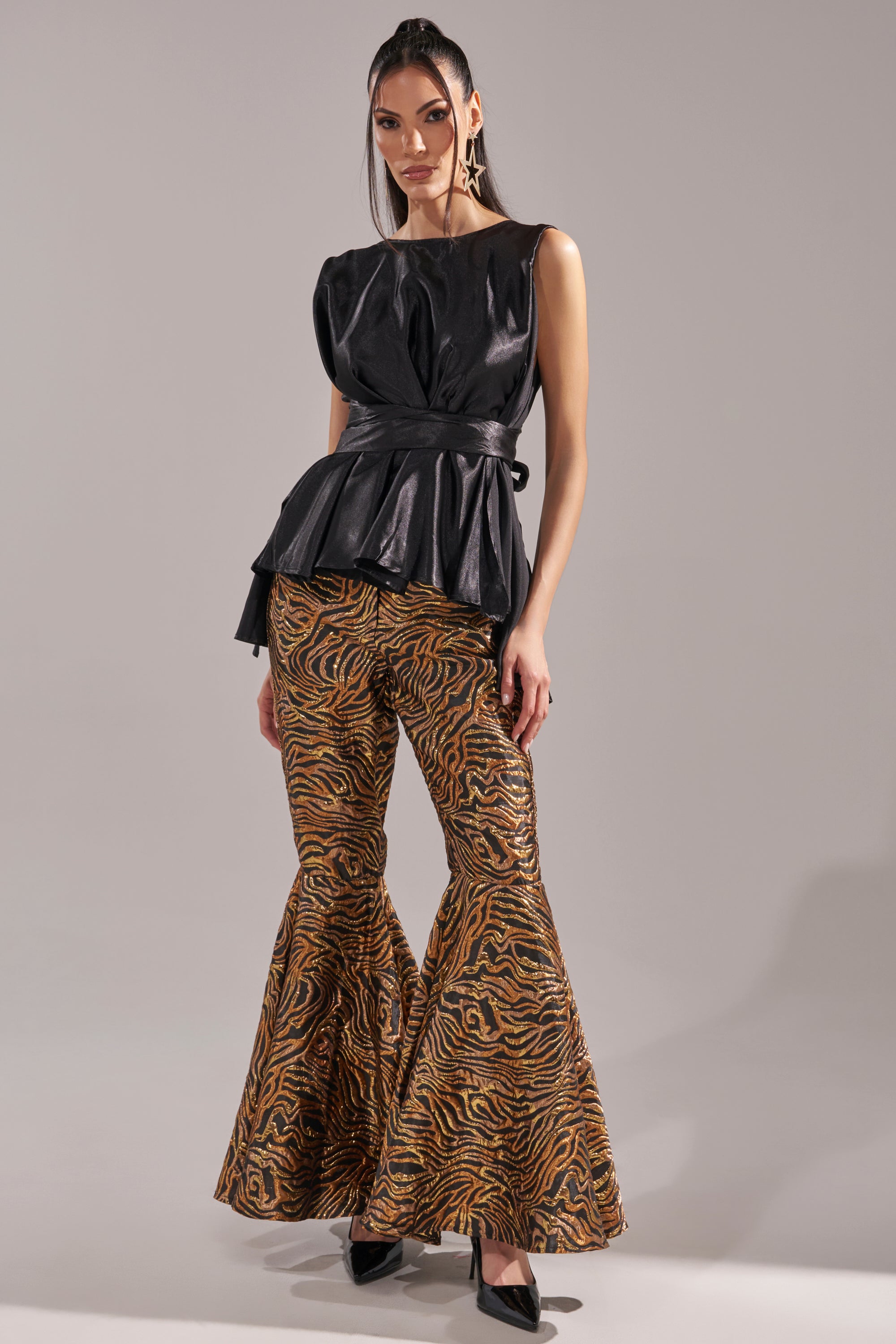 A woman poses in GET IN MOTION BROCADE FLARE TROUSER, paired with a sleeveless black peplum top and black heels. She has long, straight hair and stands against a plain gray background. 0