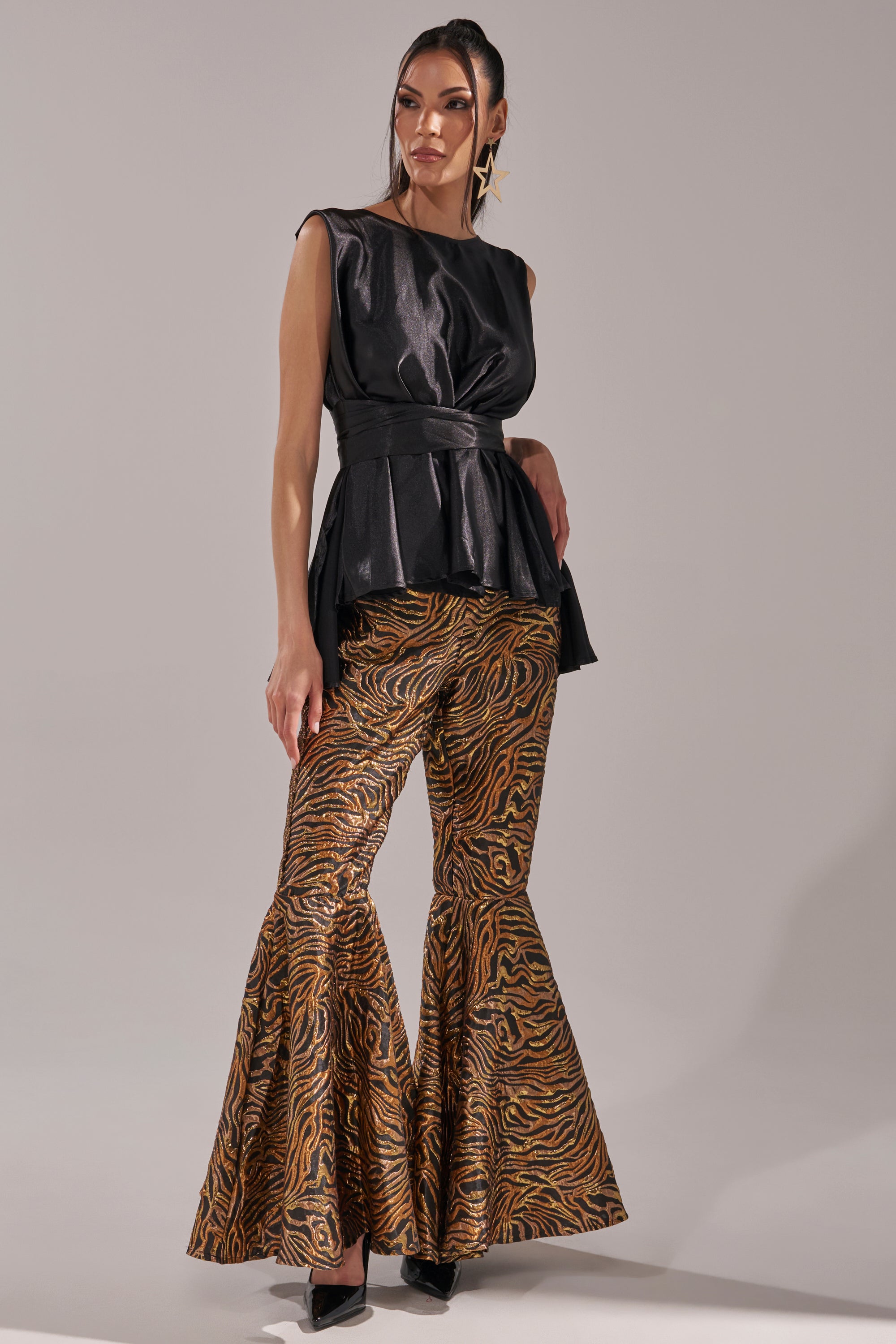A woman stands confidently in the GET IN MOTION BROCADE FLARE TROUSER, featuring a gold and brown tiger print, paired with a sleeveless black belted top and black heels. Her hair is styled in a sleek ponytail. 6