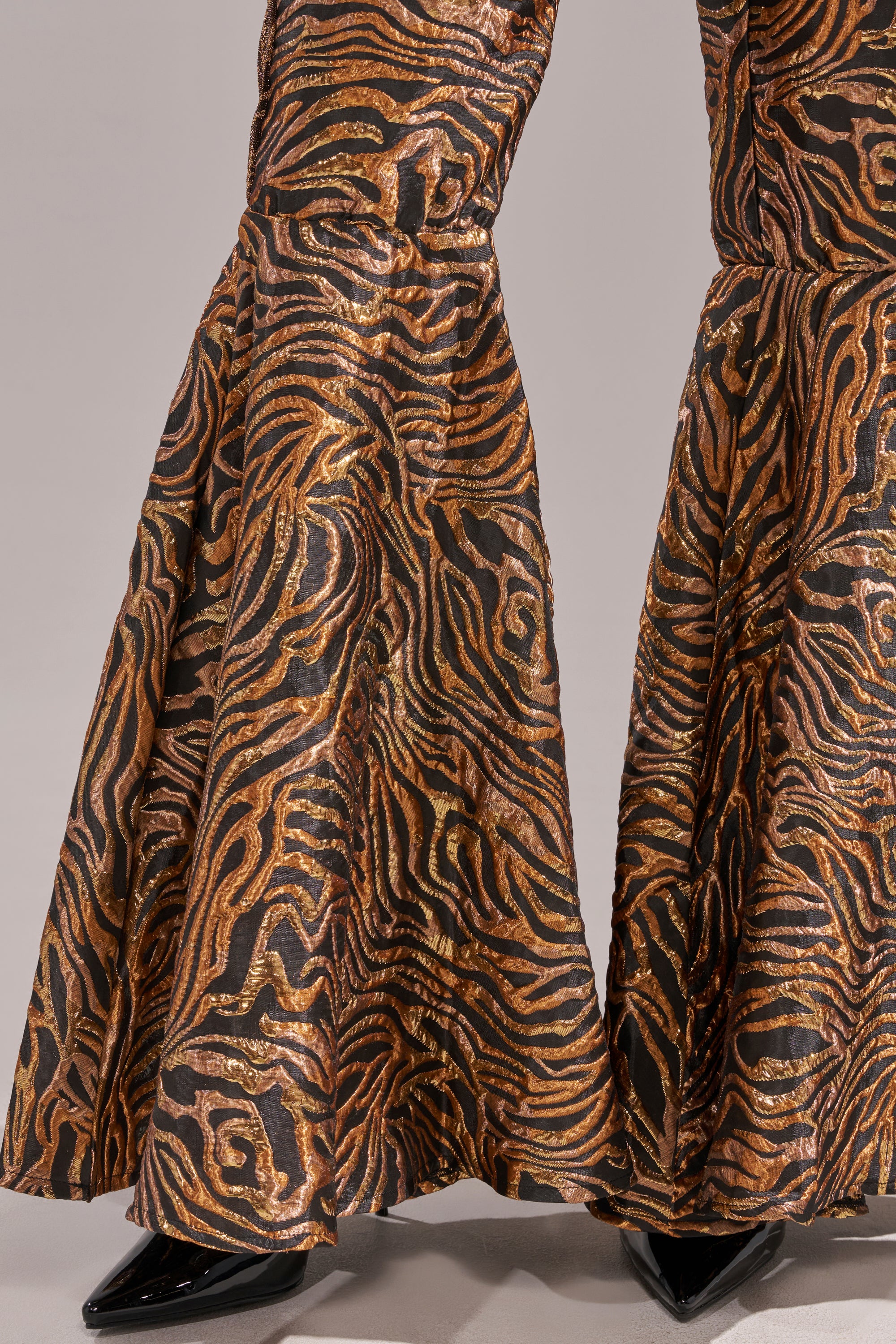 A close-up of the GET IN MOTION BROCADE FLARE TROUSER, featuring a shiny metallic jacquard with tiger-stripe pattern in orange, black, and gold. The high-rise bell flare cut is styled with pointed black shoes on a light gray background. 8