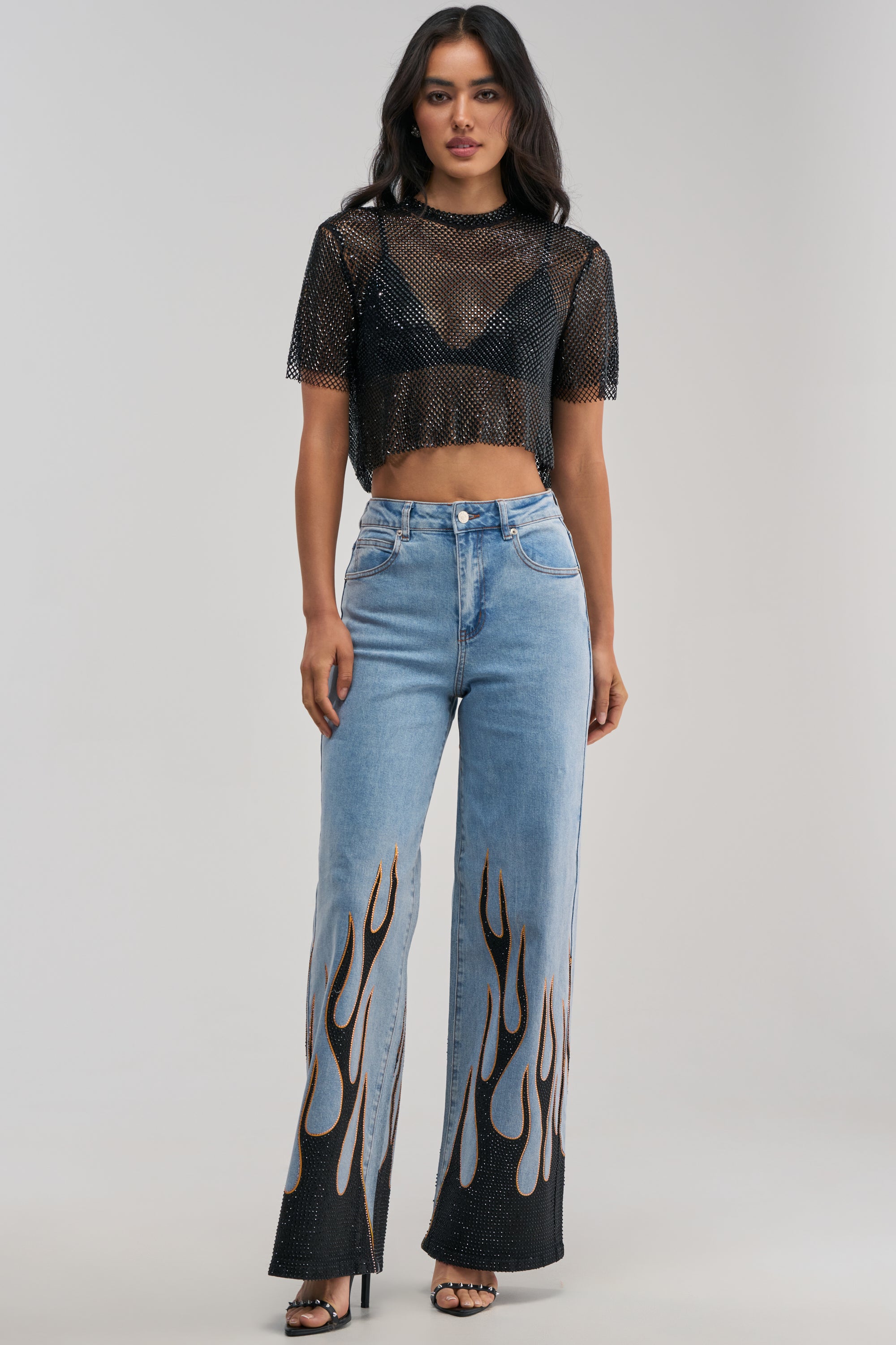 A woman showcases streetwear flair against a minimalist backdrop, wearing ON FIRE RELAXED DENIM—light blue high-waisted, wide-leg jeans featuring bold black flame designs and rhinestone fire detailing on the legs—paired with a black mesh crop top over a bralette and black heeled sandals.