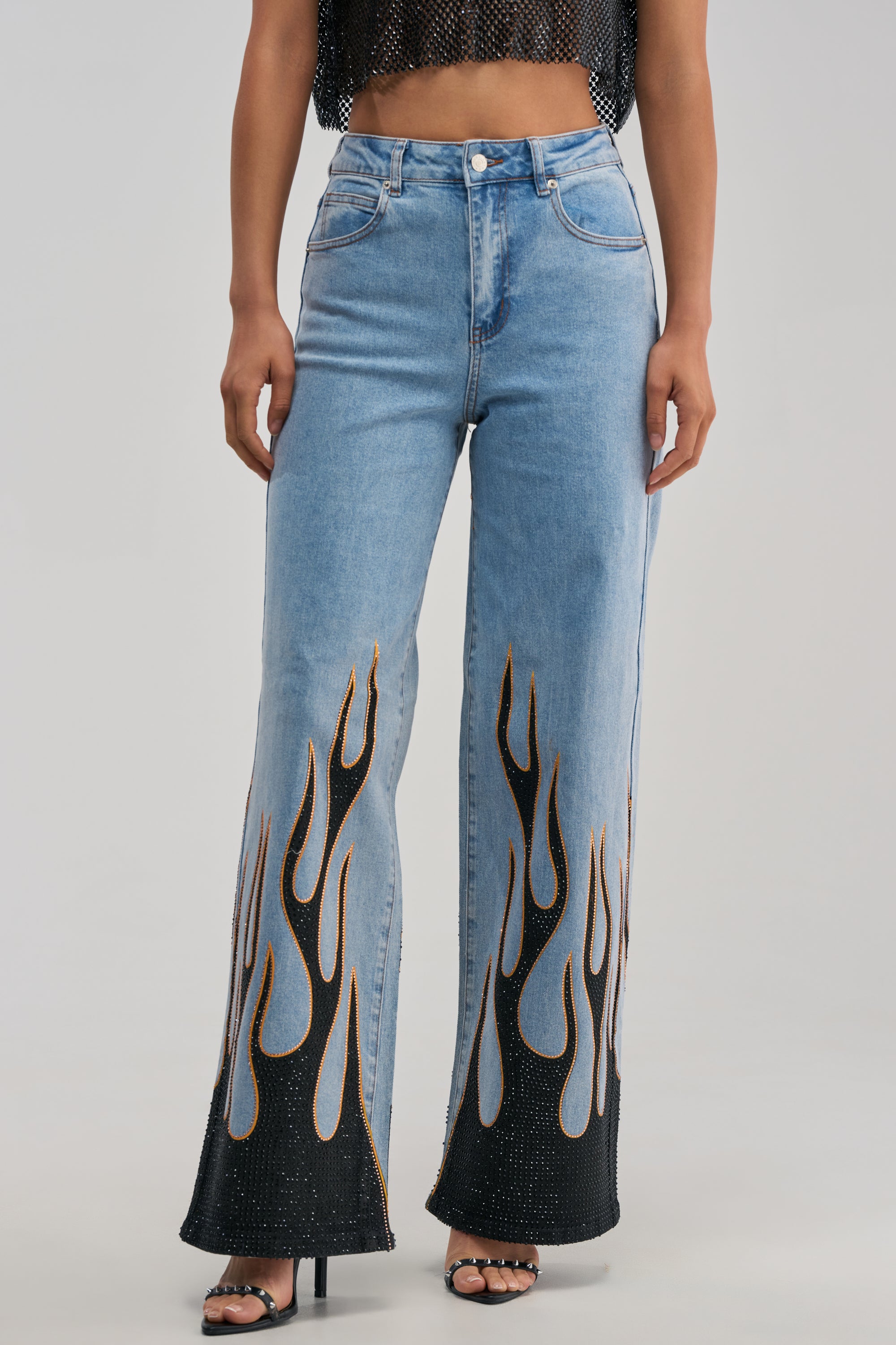A person wears the ON FIRE RELAXED DENIM—high-waisted, light blue wide-leg jeans featuring striking black and orange flame designs on the lower legs—paired with black heeled sandals and a cropped, sleeveless black top for a bold streetwear look.