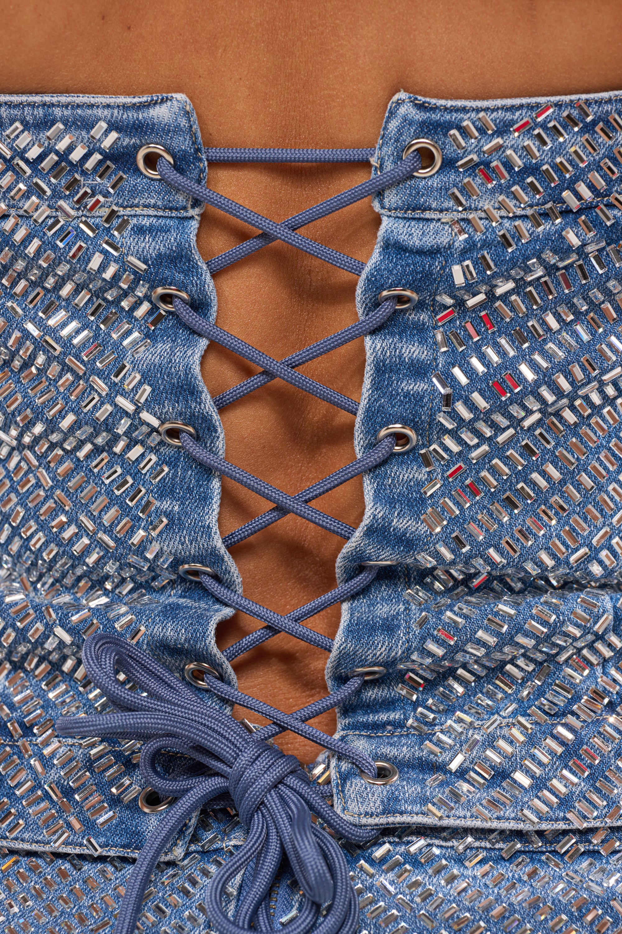 Close-up of the BLING BLING CORSET, a rhinestone denim piece with silver accents, featuring blue cords laced up the back and crisscrossed to reveal some skin. 11
