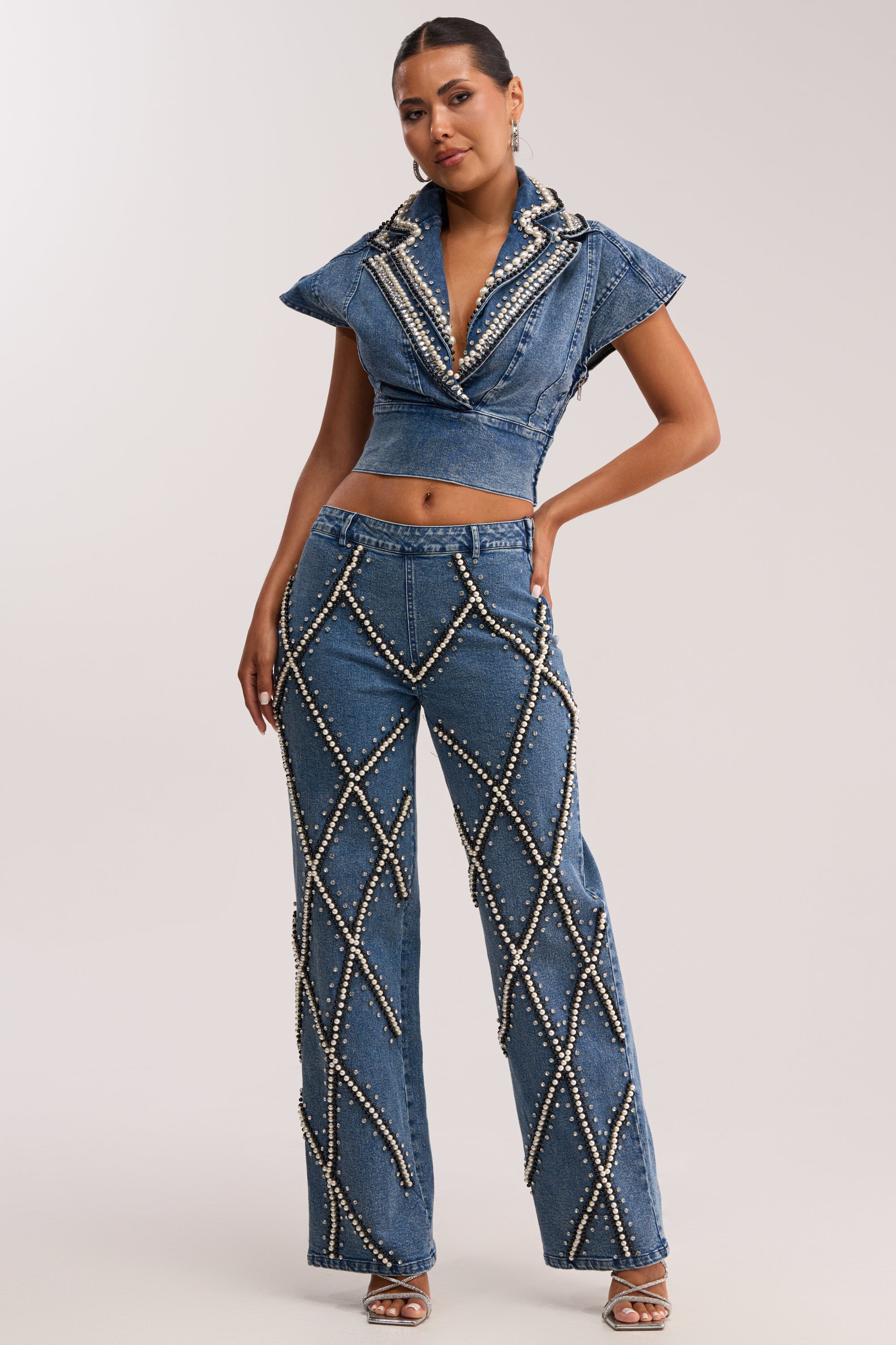 A woman models the DRIPPING IN PEARLS DENIM BLOUSE, a high-collared, short-sleeve denim top adorned with pearl crisscross patterns, paired with matching flared jeans for a chic streetwear look.