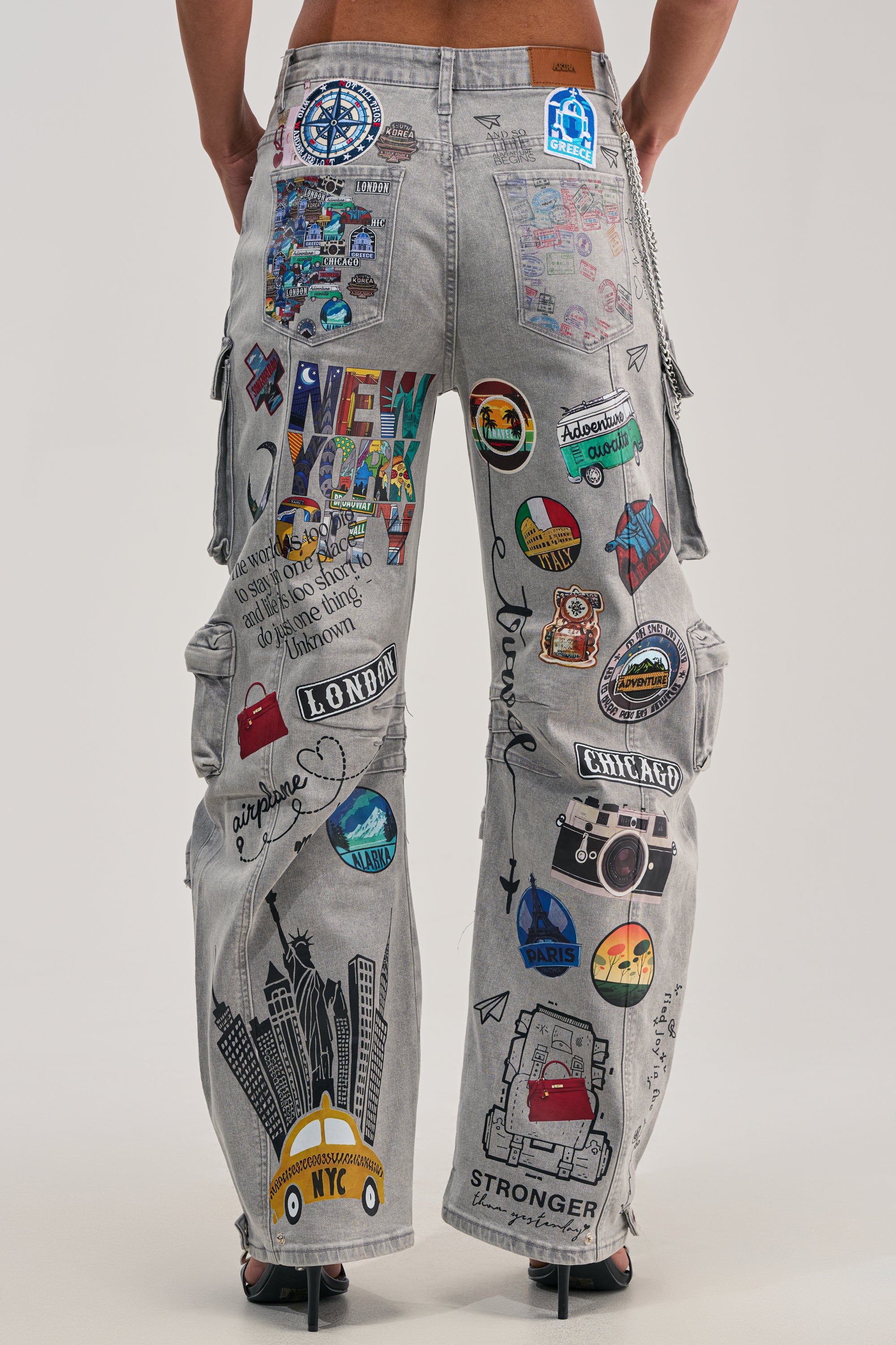 Seen from the back, a person wears RAJ CARGO DENIM: grey denim cargo pants adorned with vibrant graphic patches and travel-inspired artwork of cities like New York, London, Chicago, and Paris—featuring taxis, cameras, and streetwear badges.