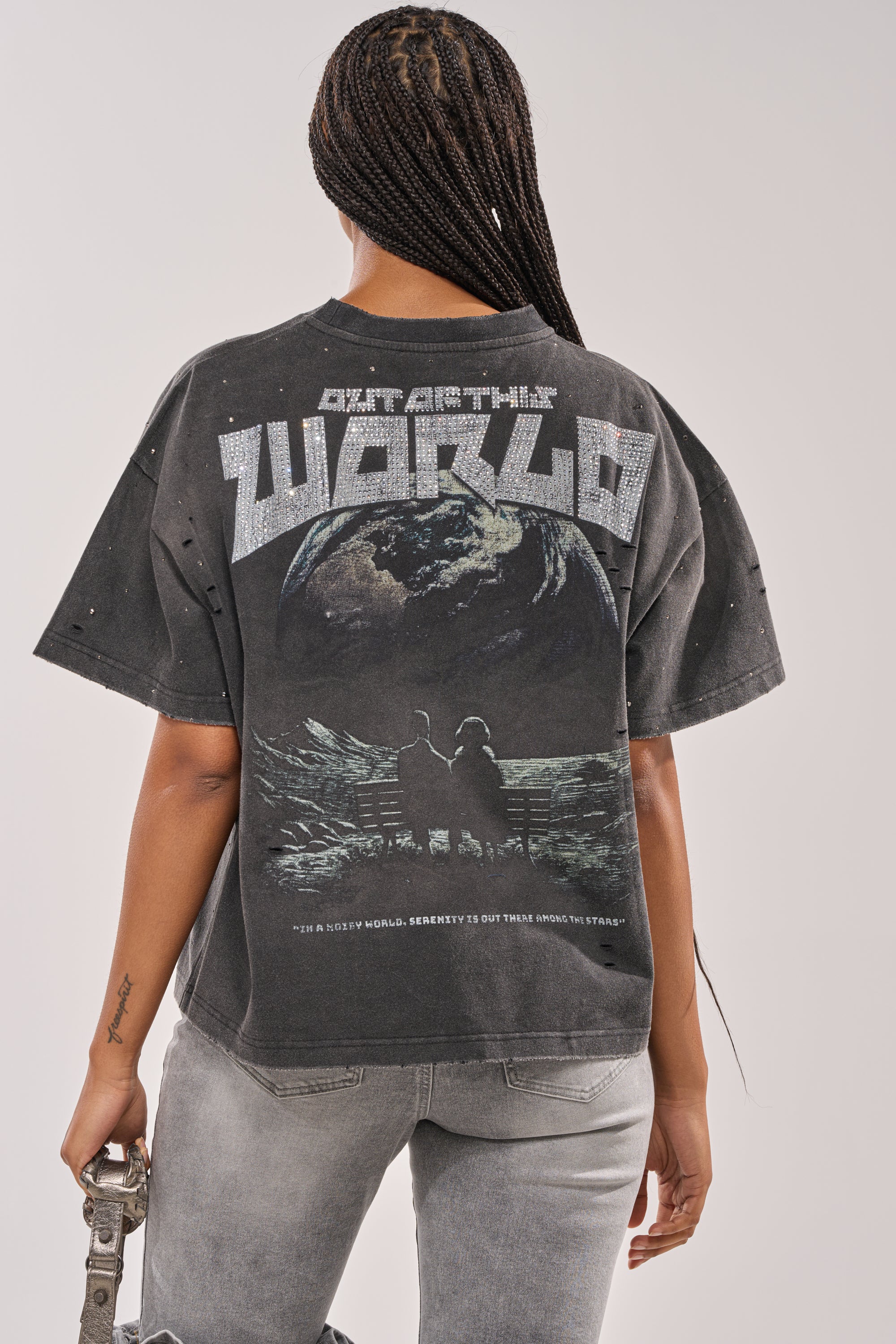 A person with braided hair, seen from behind, wears the OUT OF THIS WORLD GRAPHIC TEE featuring two people on a bench gazing at Earth from space, with "OUT OF THIS WORLD" above the graphic. 3