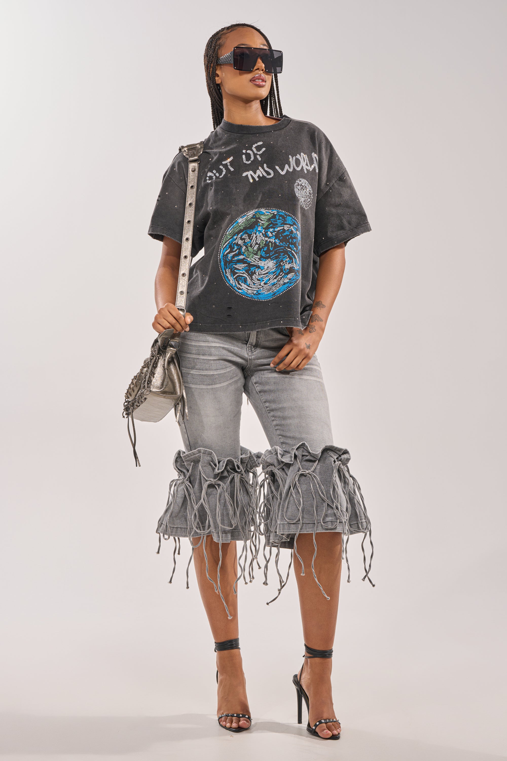 A woman stands confidently against a plain background wearing sunglasses, high heels, a textured shoulder bag, distressed gray denim shorts with frayed fringe, and the OUT OF THIS WORLD GRAPHIC TEE. 4