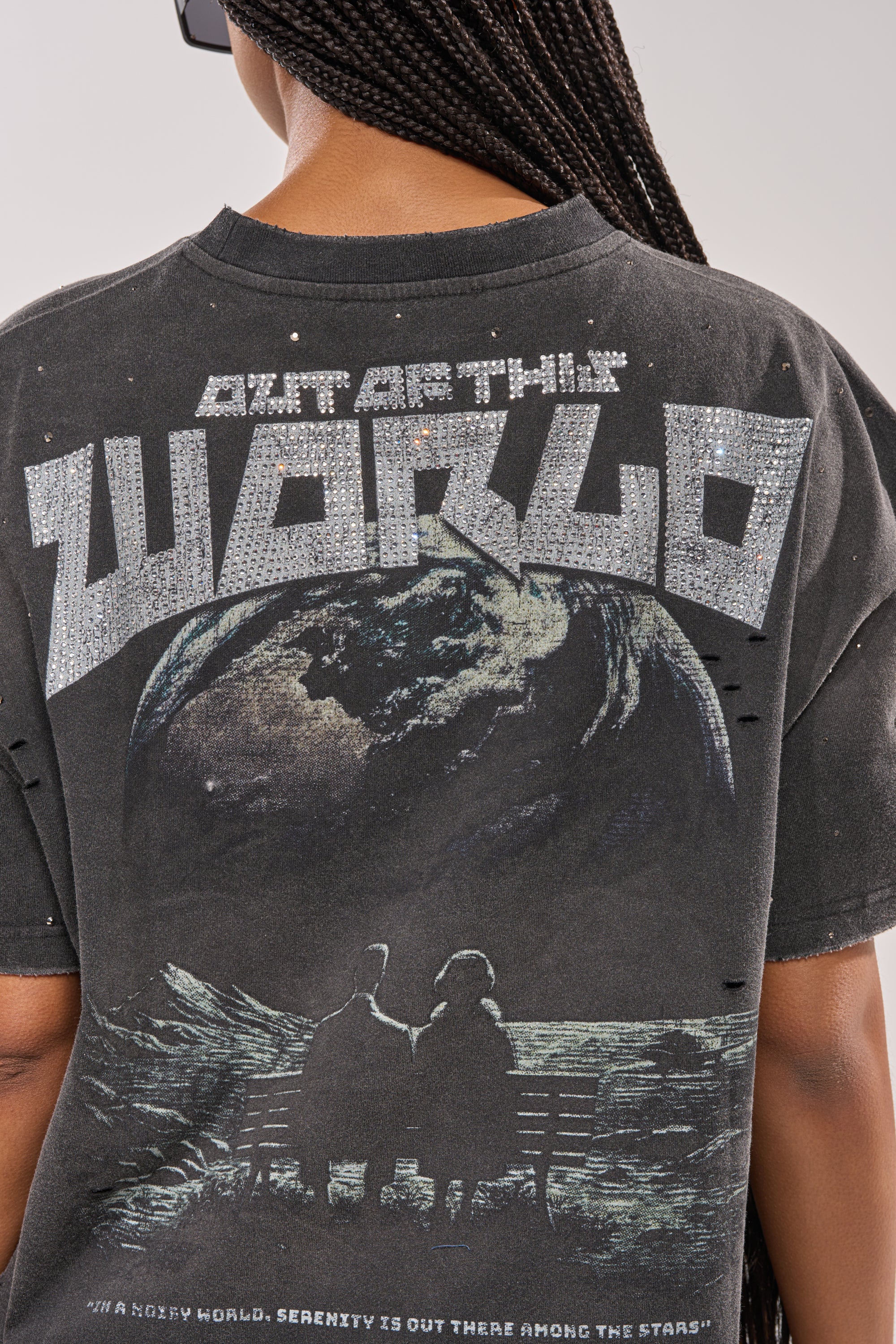 A person with braided hair wears the OUT OF THIS WORLD GRAPHIC TEE—an oversized streetwear shirt with two people on a bench gazing at Earth from space, and “OUT OF THIS WORLD” in glittery letters above the cosmic scene. 5